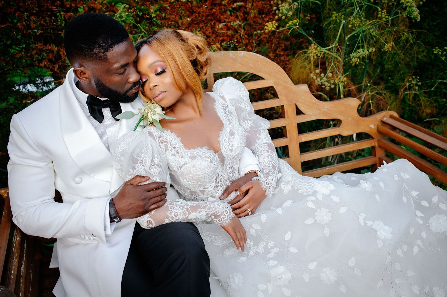 Wedding couple in white attire sharing an intimate moment on a garden bench.