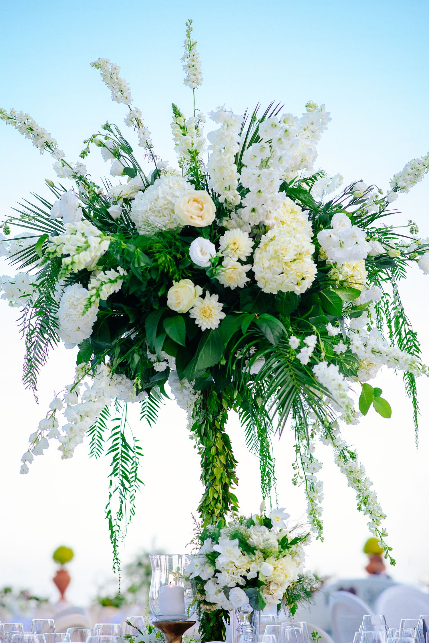 Elegant white floral wedding arch arrangement with roses and greenery against blue sky.
