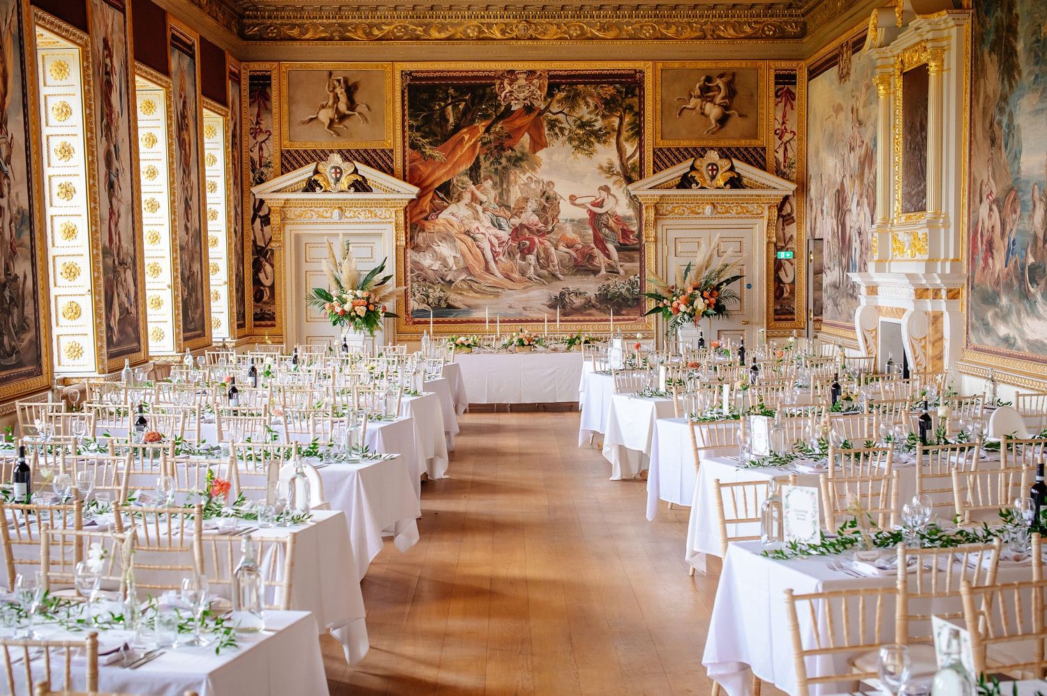 Elegant wedding reception setup in ornate historic hall with white tables and gold chairs beneath classical painted murals.