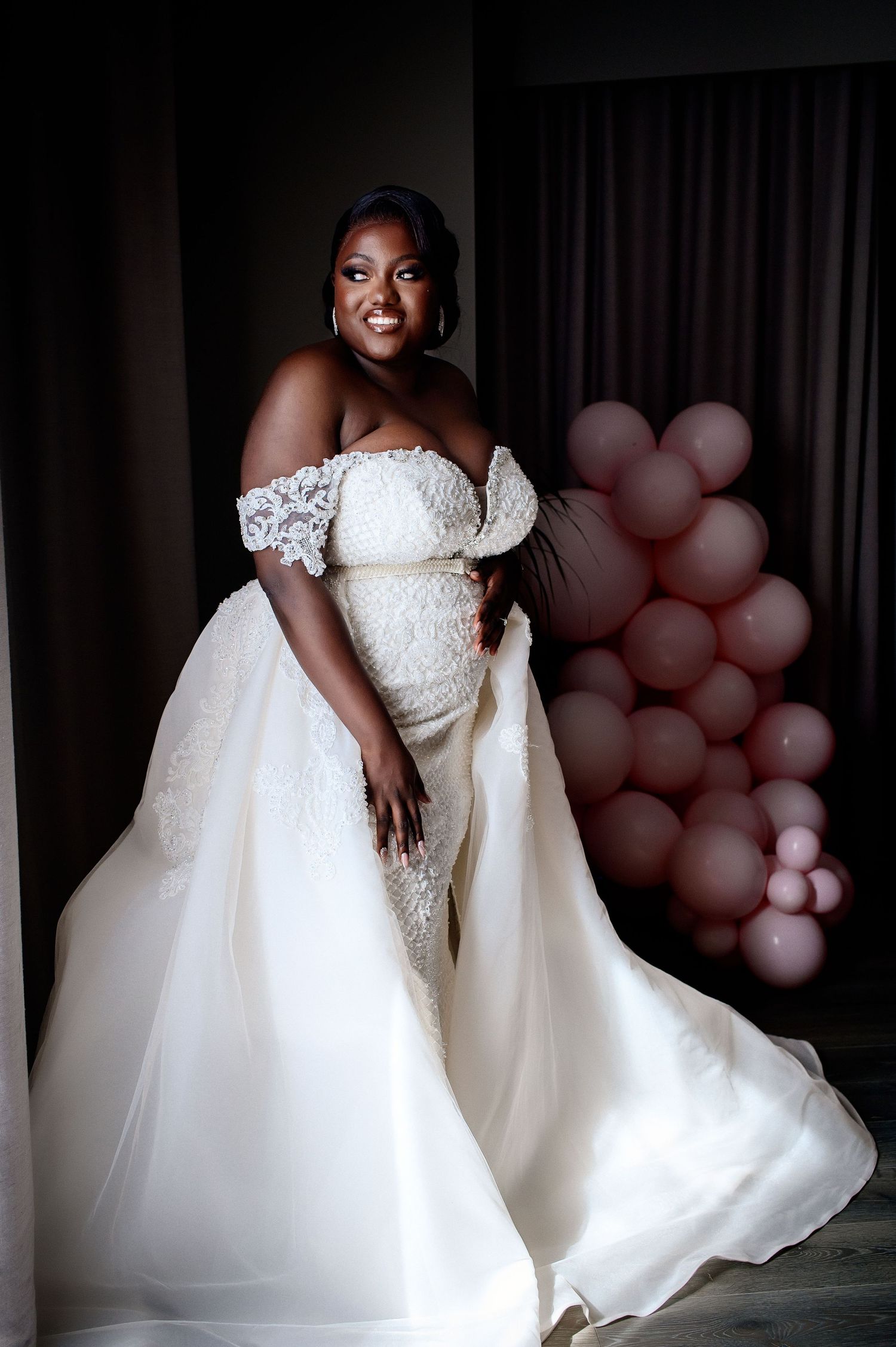 Elegant wedding dress with off-shoulder design and pink balloon decor in background.