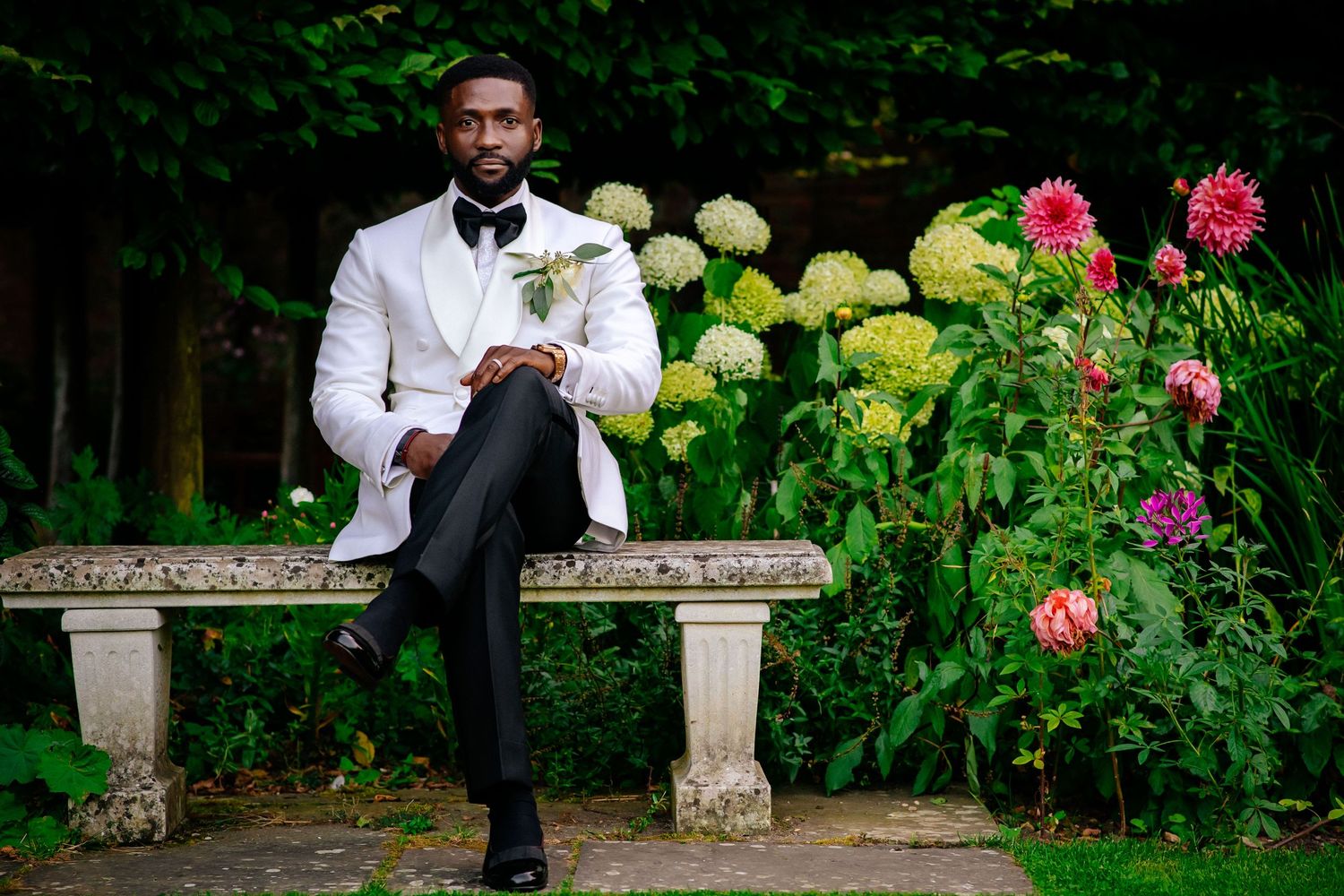 Person in white suit sitting on stone bench beside colorful garden flowers.