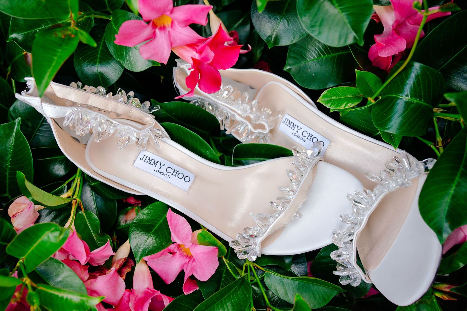 Elegant white bridal shoes with crystal embellishments among pink flowers and green leaves.