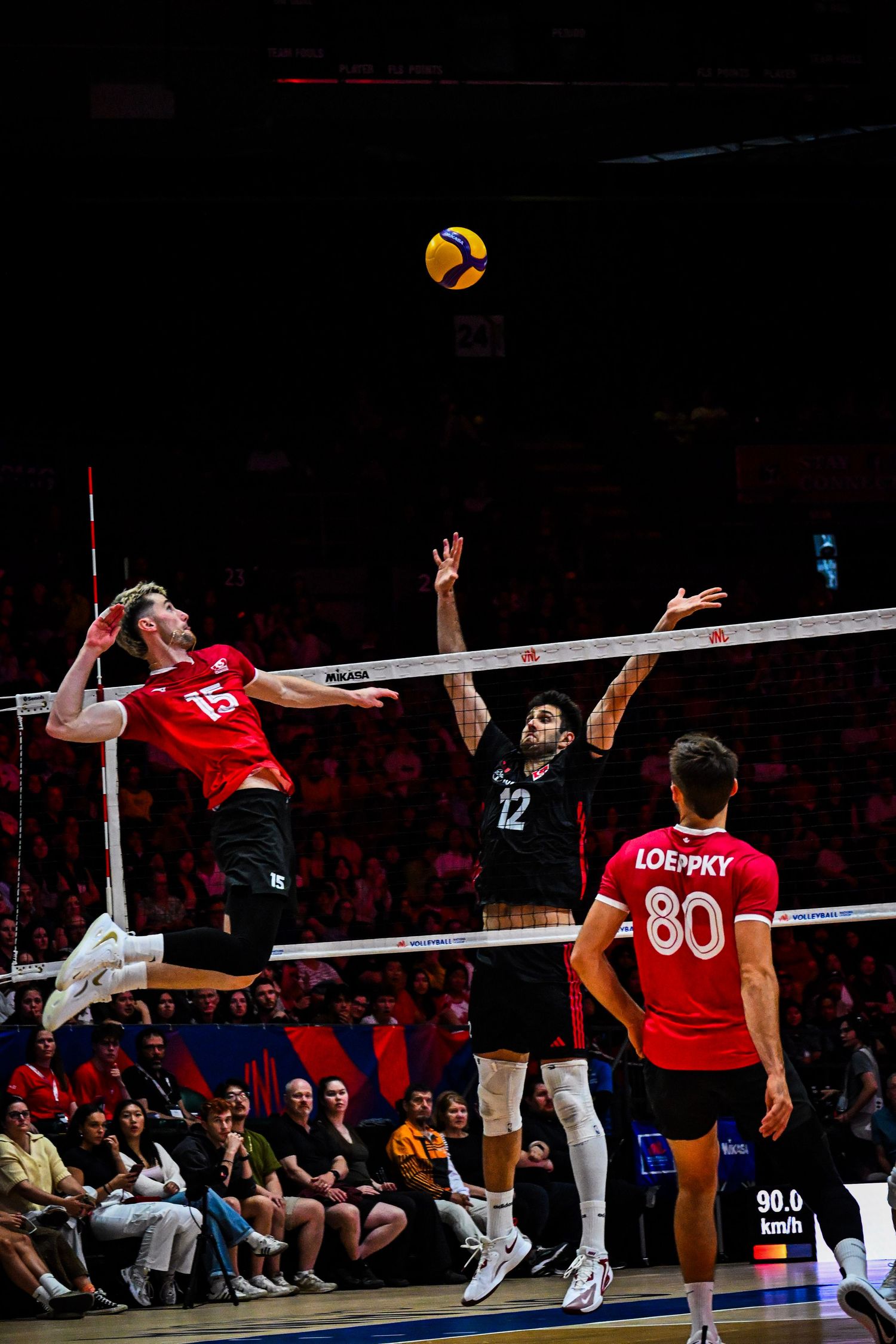 Volleyball player in a red jersey leaps high to spike the ball over an opposing defender at the net.