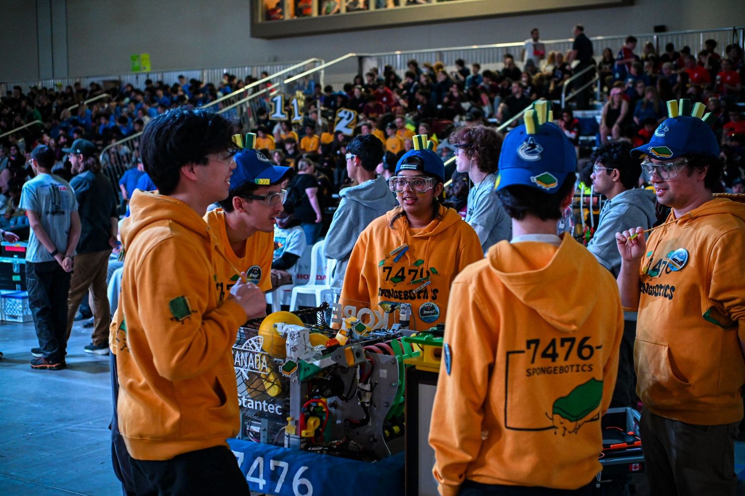 Robotics team members in matching yellow hoodies collaborating in a packed tournament stadium.
