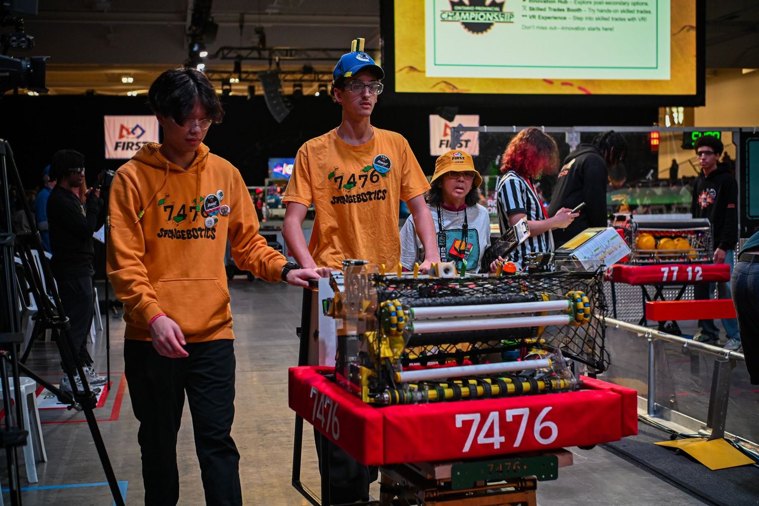 Team members standing beside competition robot 7476 in an indoor arena setting.