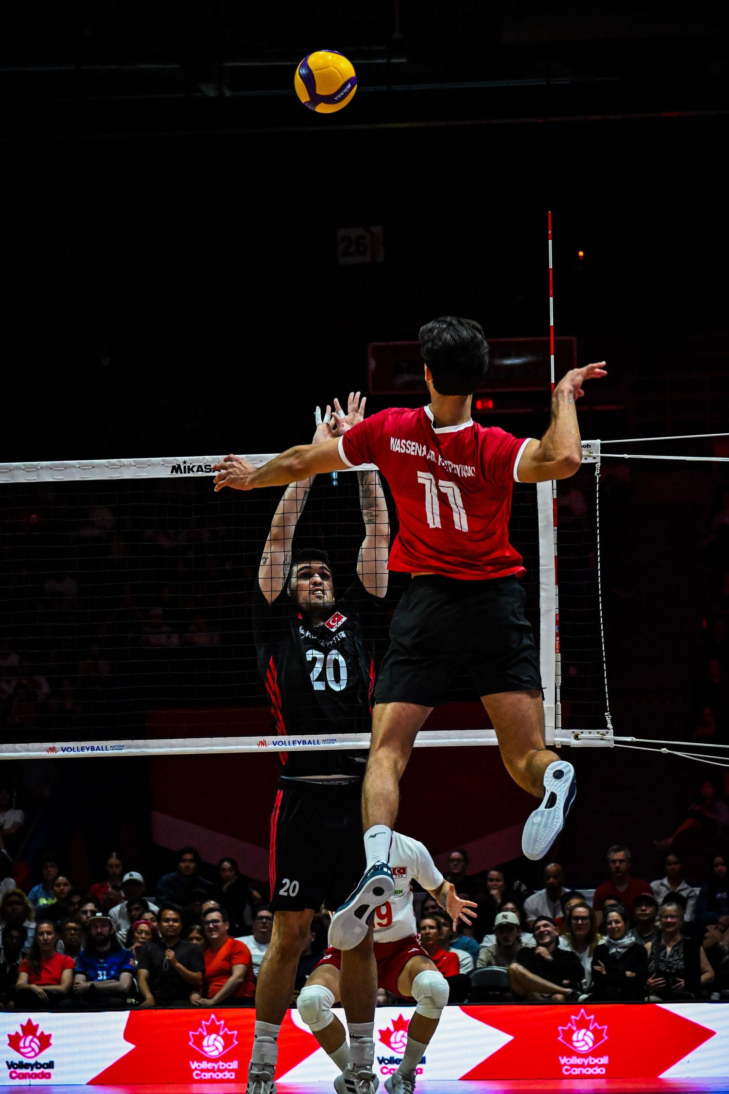 Volleyball player in a red jersey jumps high in the air to spike a ball above the net during an indoor match.