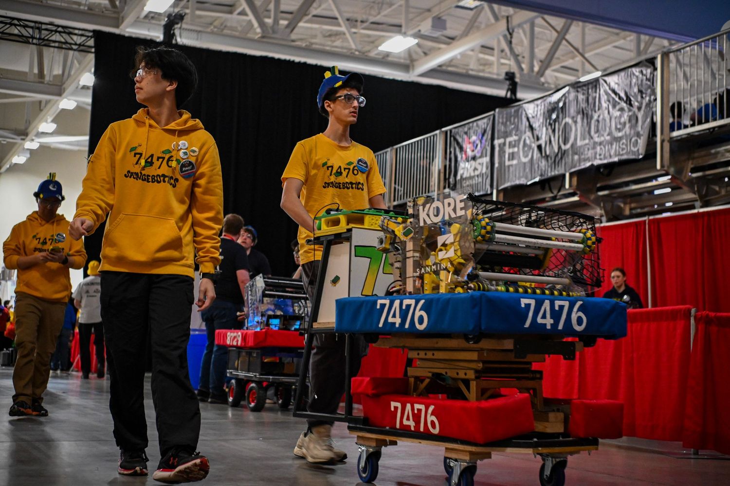 Team members walking past a robotics pit station in a large indoor competition arena.