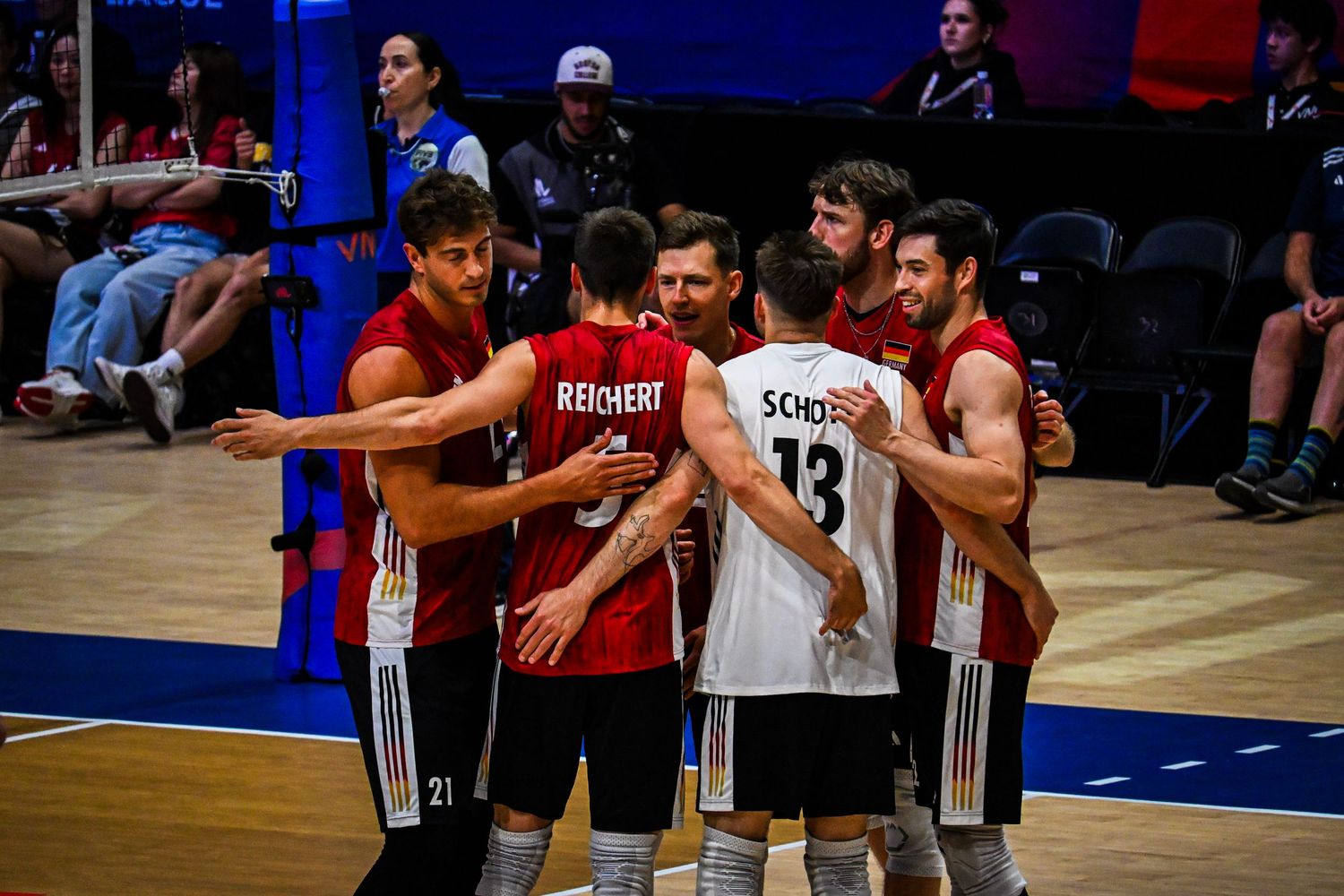 A volleyball team huddling together on the court during an indoor game with spectators in the background.