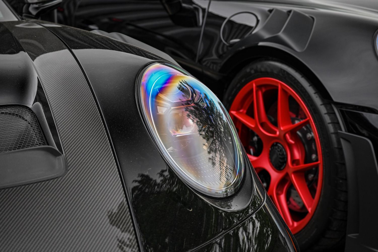 Extreme close-up of a glossy black sports car's circular headlight reflecting colorful lights next to a bright red multi-spoke wheel.