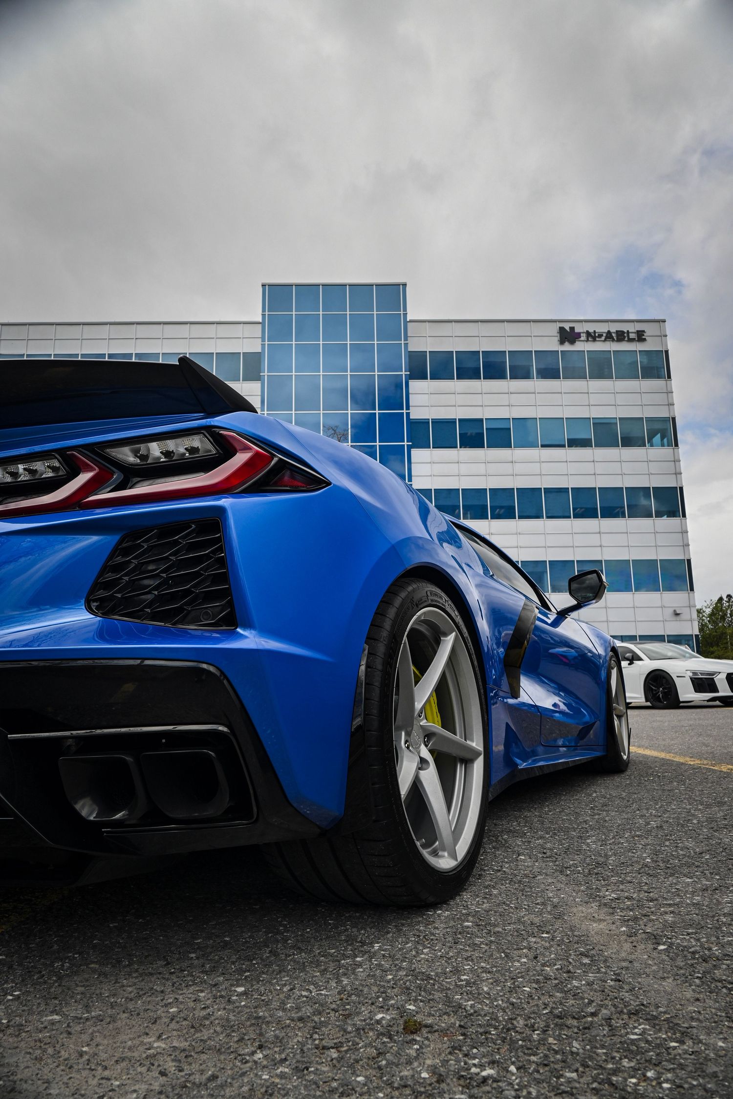 Vertical close-up view from behind the rear wheel of a blue modern Chevrolet Corvette, showing its sharp angular body line and taillight.
