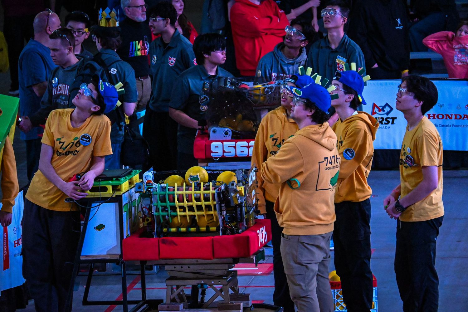 A group of robotics team members in yellow shirts gathered around their competition robot in a crowded venue.
