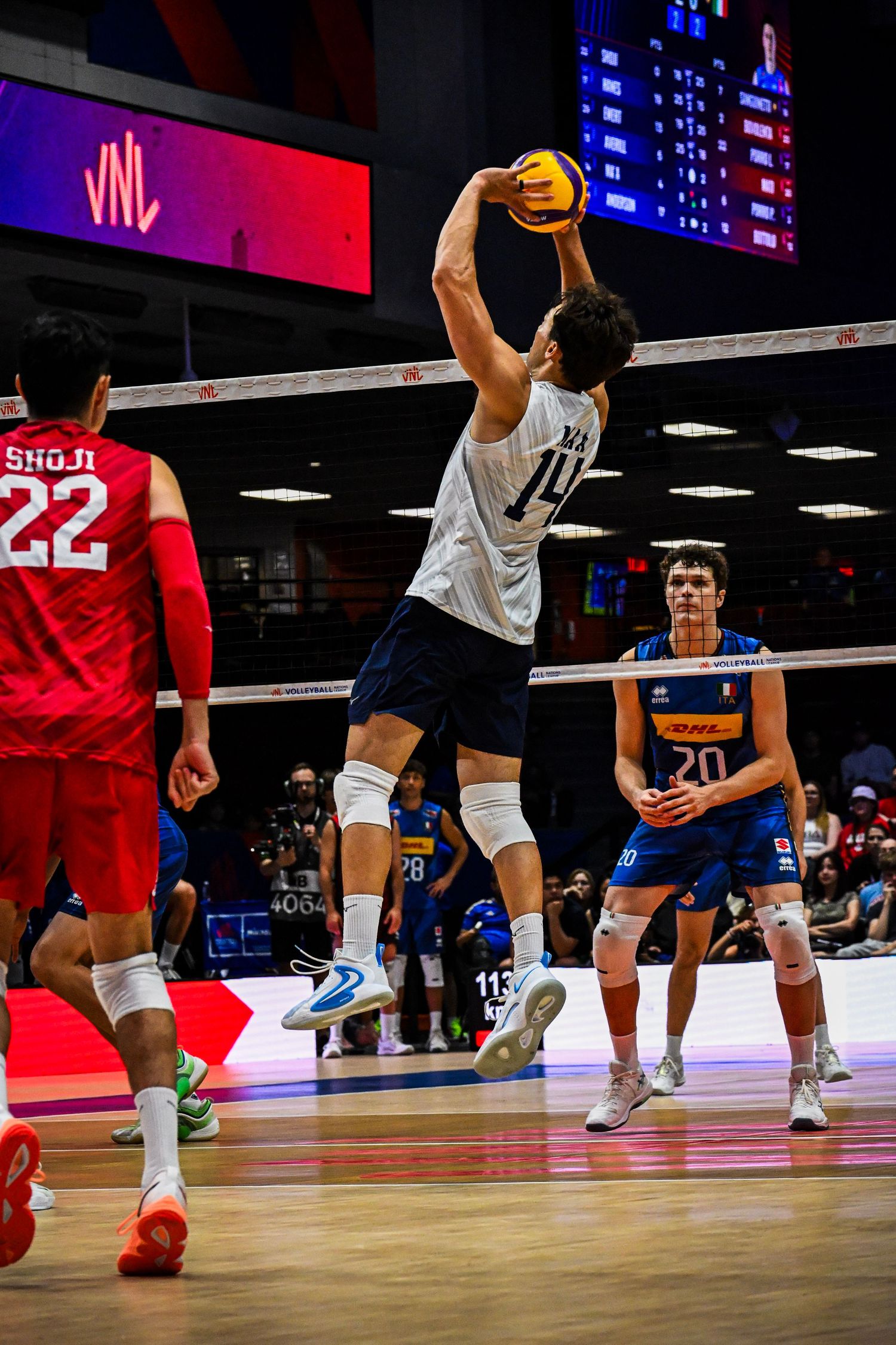 Volleyball player in a white jersey jumps high at the net to block an opponent's shot during a match.