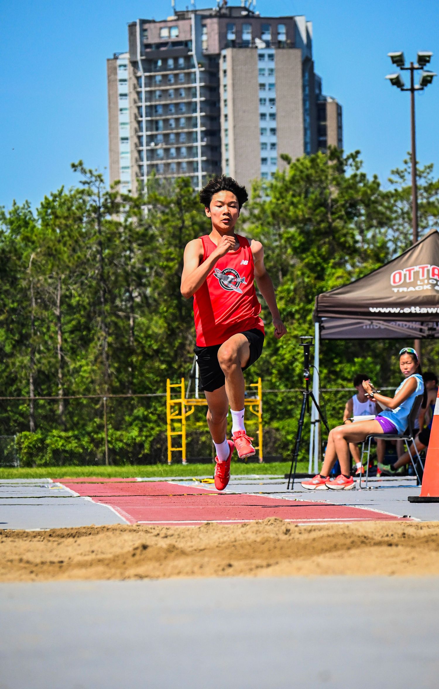 An athlete in a red jersey sprinting down the runway at an outdoor track and field event.