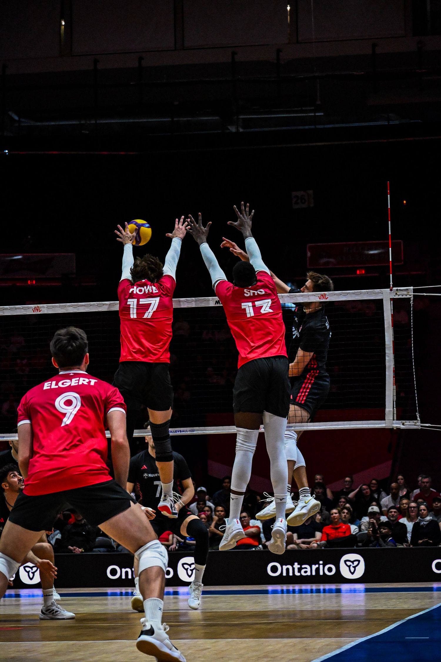 Two volleyball players in red jerseys jump up at the net to block an opponent's attack.