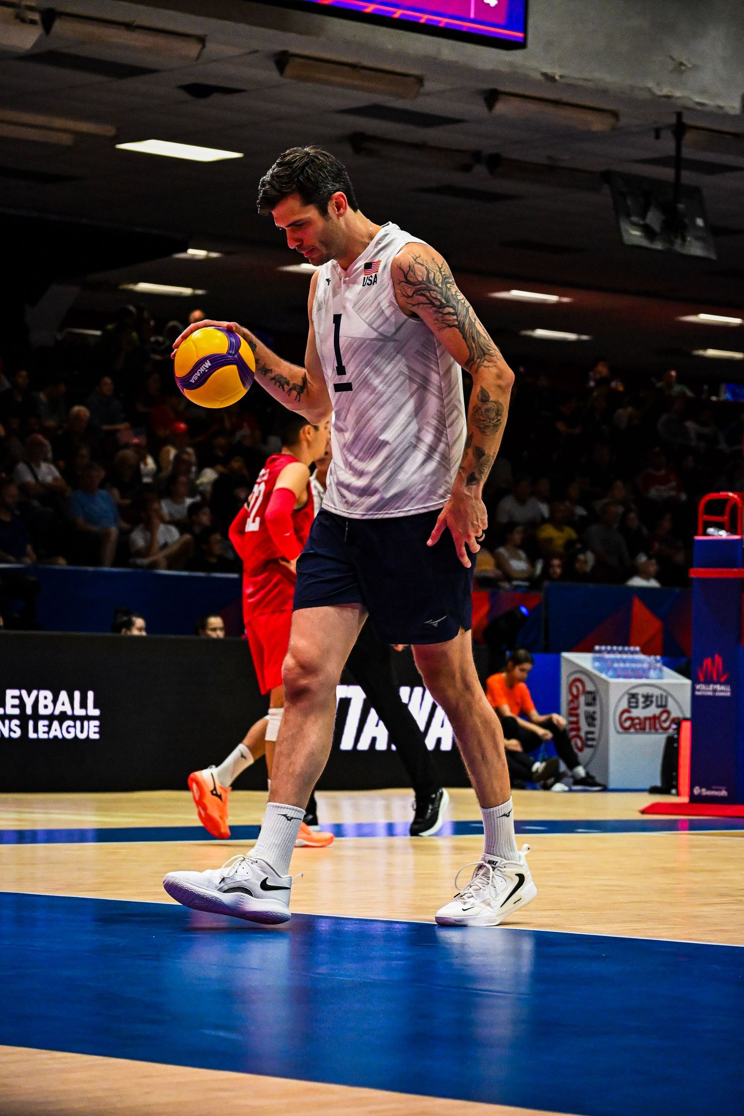 Volleyball player in a white sleeveless jersey walks across the court carrying a ball.