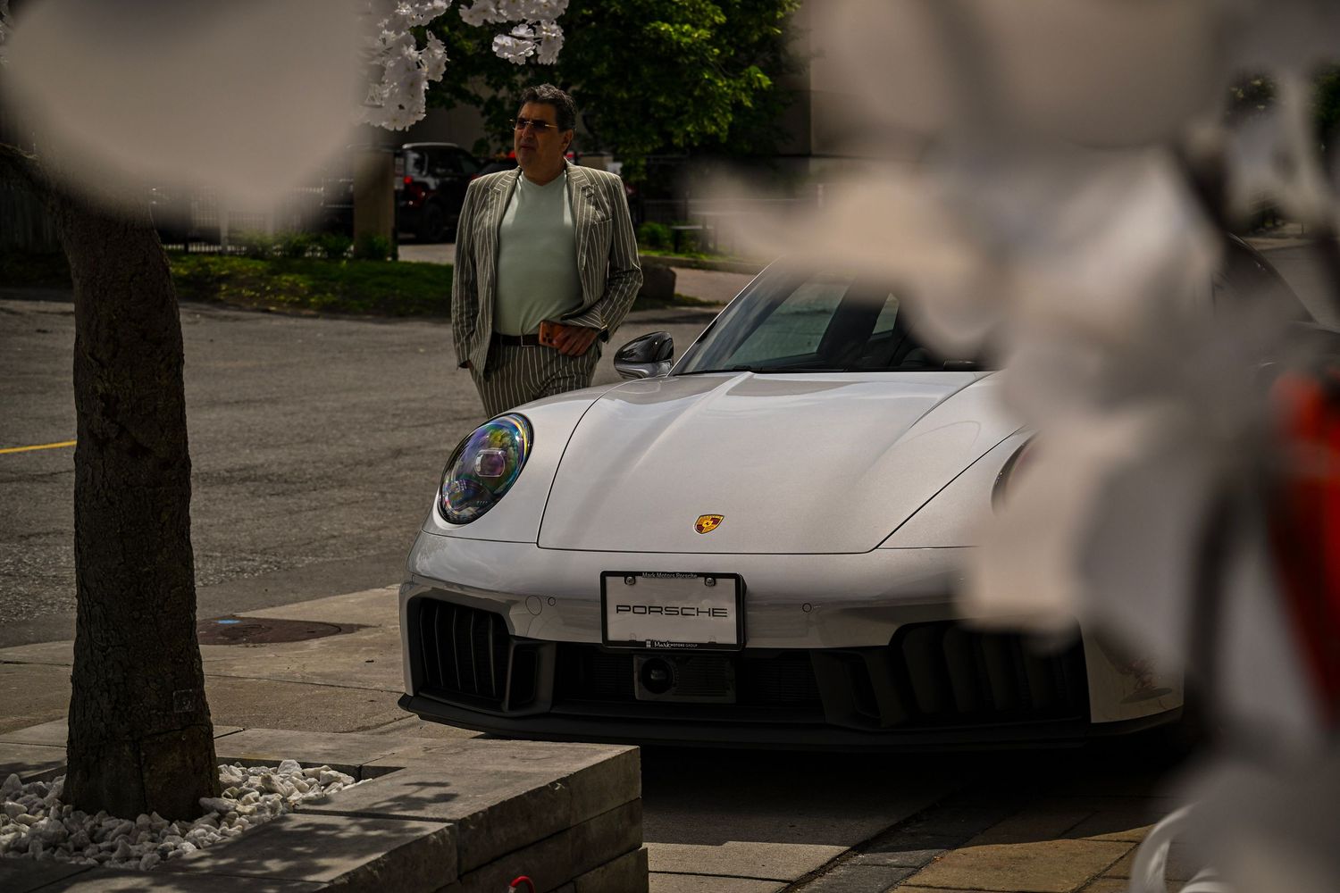 Front view of a white Porsche sports car parked on a tree-lined street with a person standing behind it.