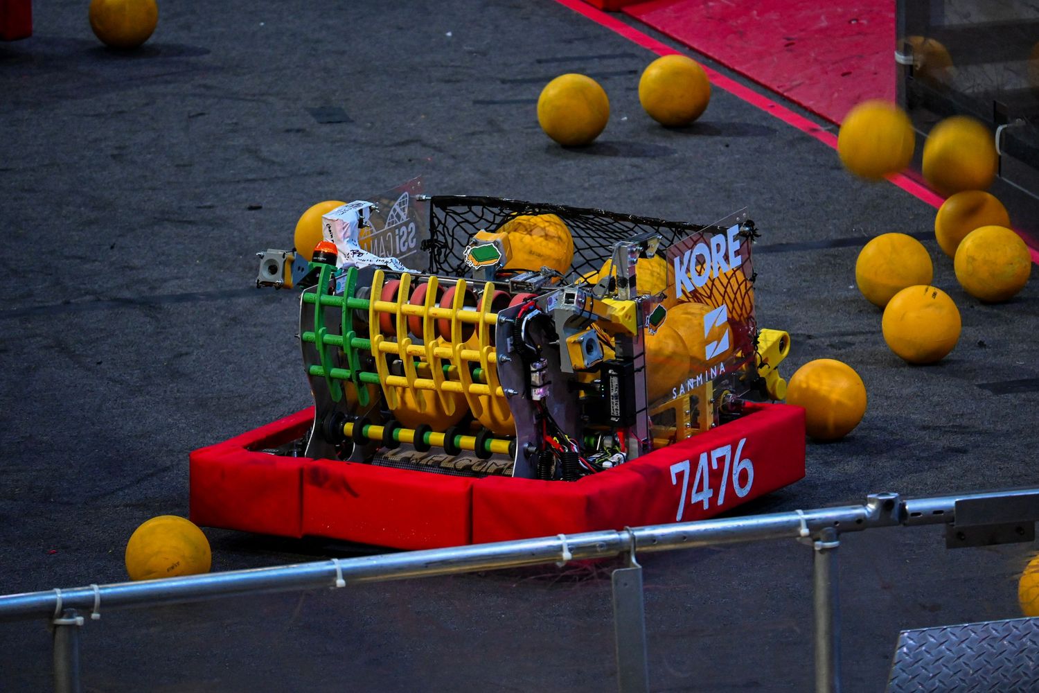 A colorful custom competition robot numbered 7476 navigating a field filled with bright yellow balls.