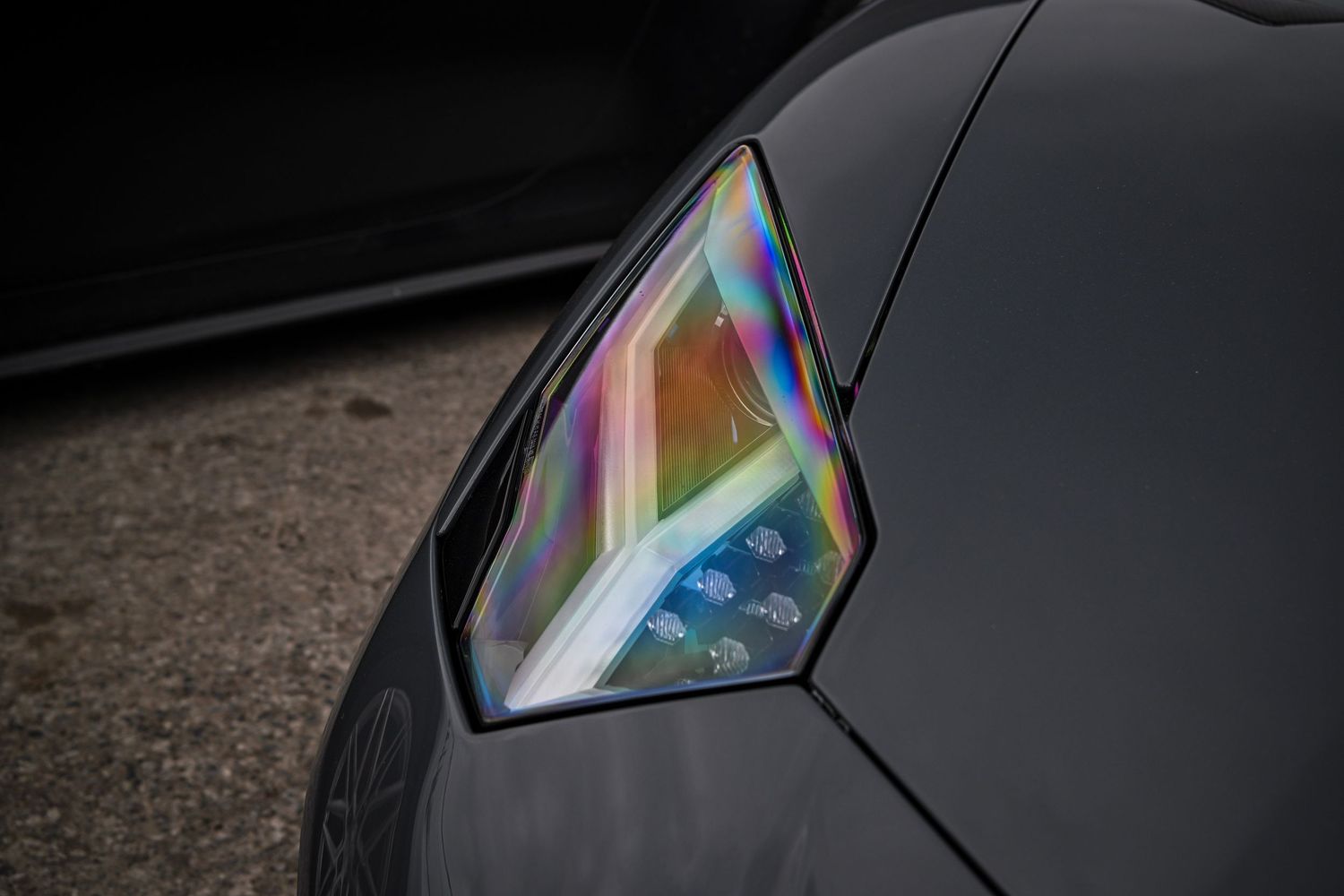 Close-up of the angular headlight of a matte black Lamborghini showing iridescent geometric interior detailing.