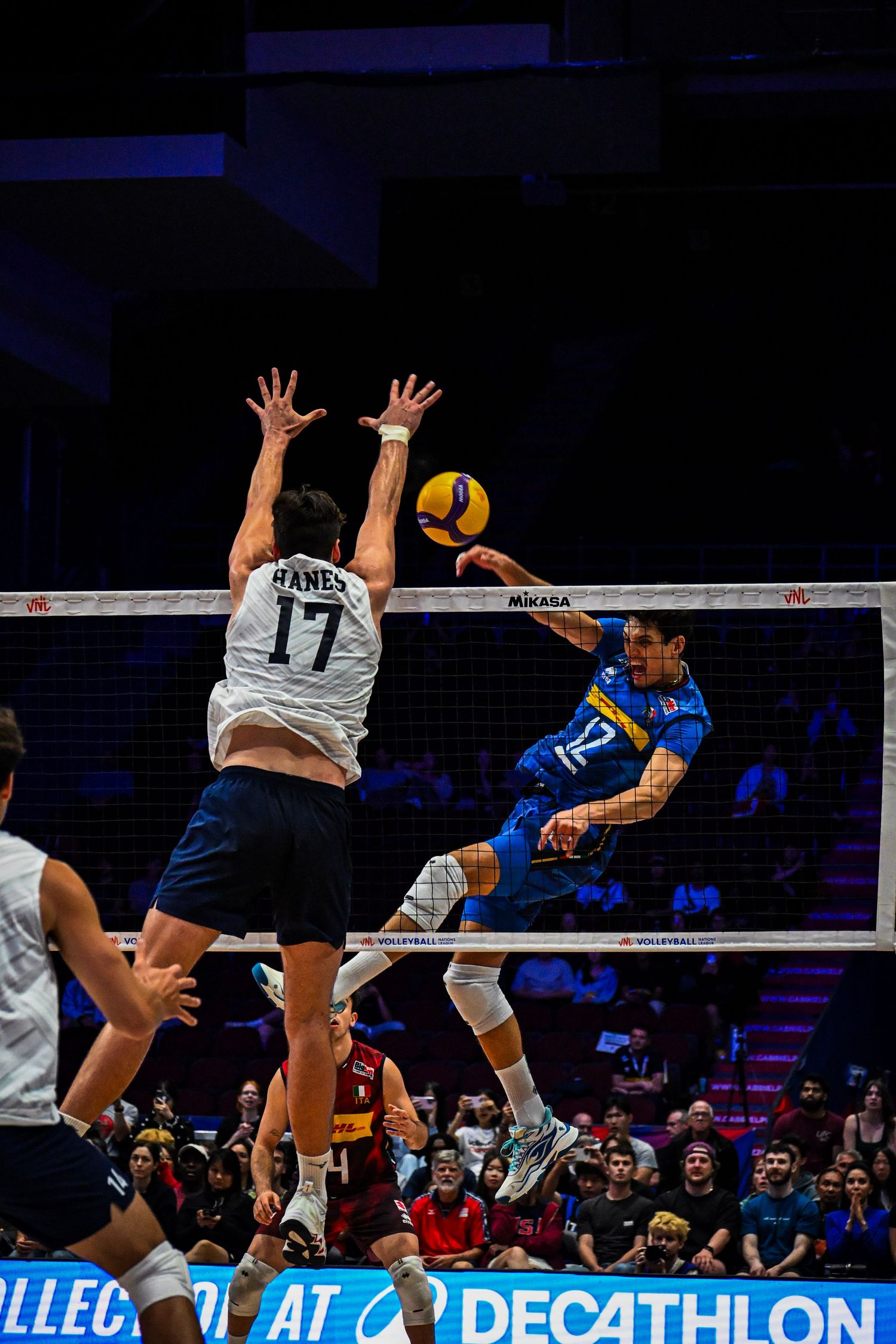 Volleyball player in blue spikes the ball over the net as defender number 17 in white leaps up to block.