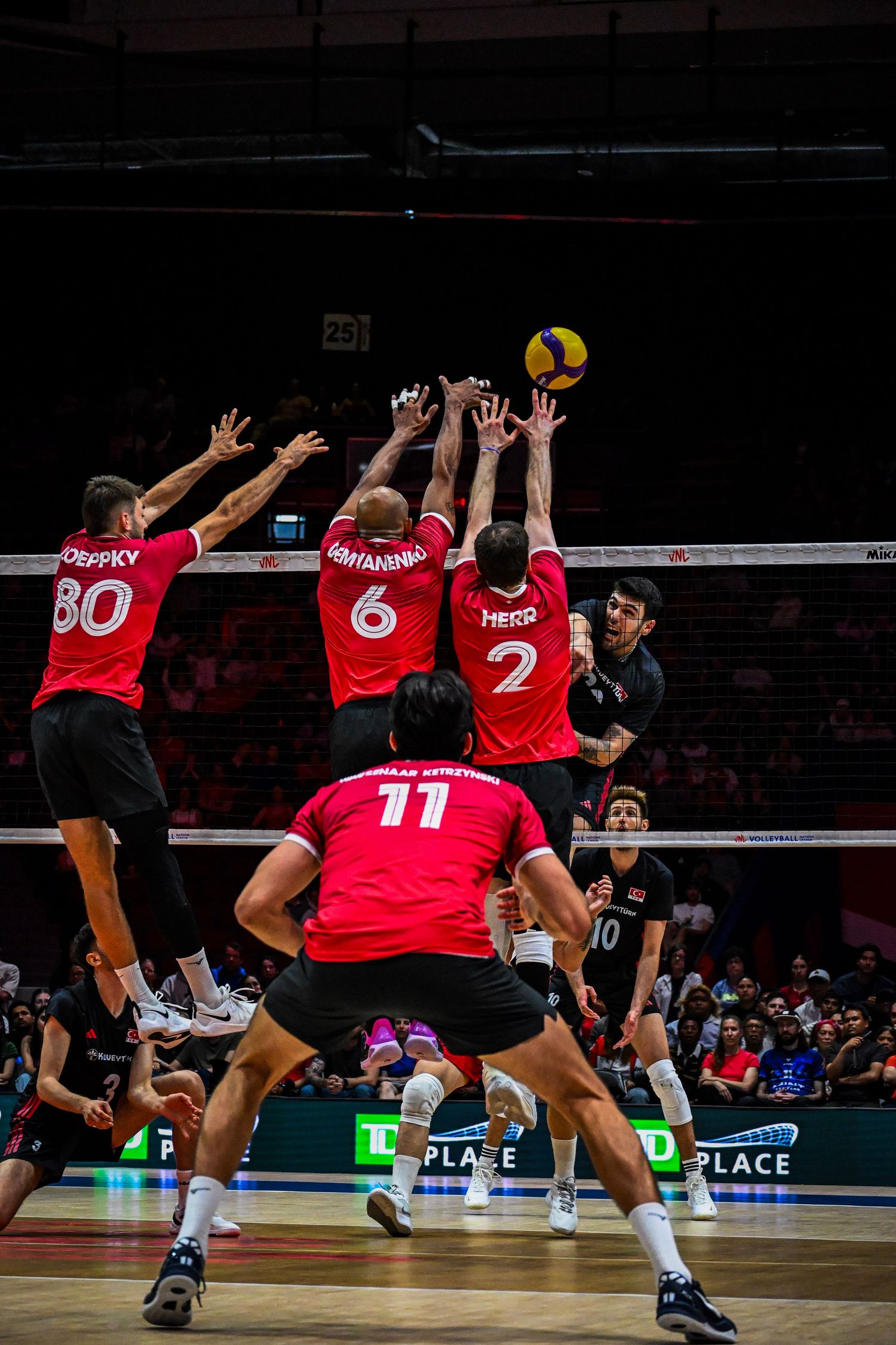 Three volleyball defenders in red jerseys leap together at the net to form a triple block.