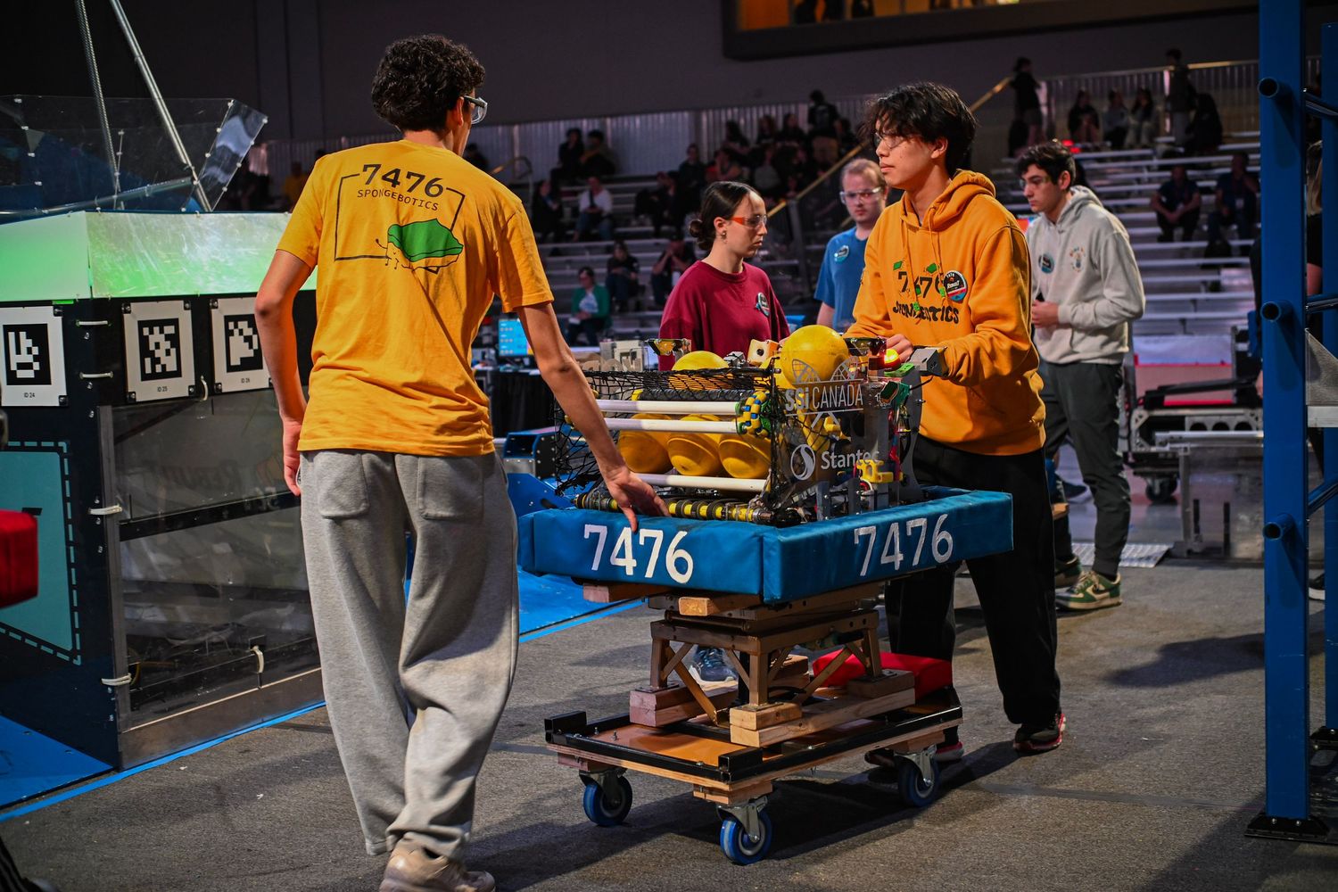 Robotics team members transporting their custom built robot on a rolling cart inside a packed stadium.