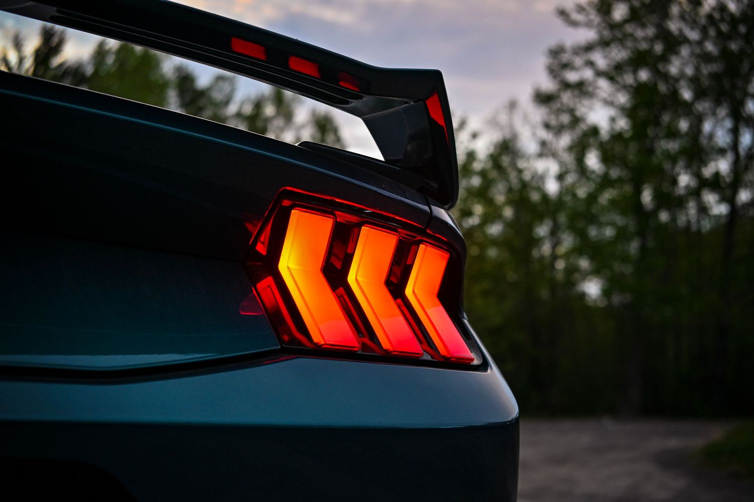Close-up of the illuminated three-bar tri-bar taillight and rear wing spoiler of a dark green Ford Mustang.