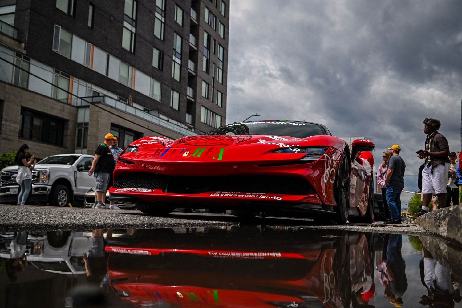 Low-angle shot of a bright red Ferrari sports car reflecting in a street puddle on a cloudy day with city buildings and onlookers.