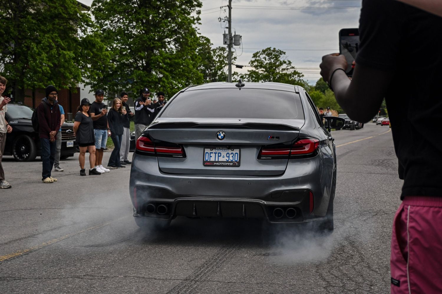 Rear view of a gray BMW sedan spinning its tires and creating smoke as crowds watch and take photos.
