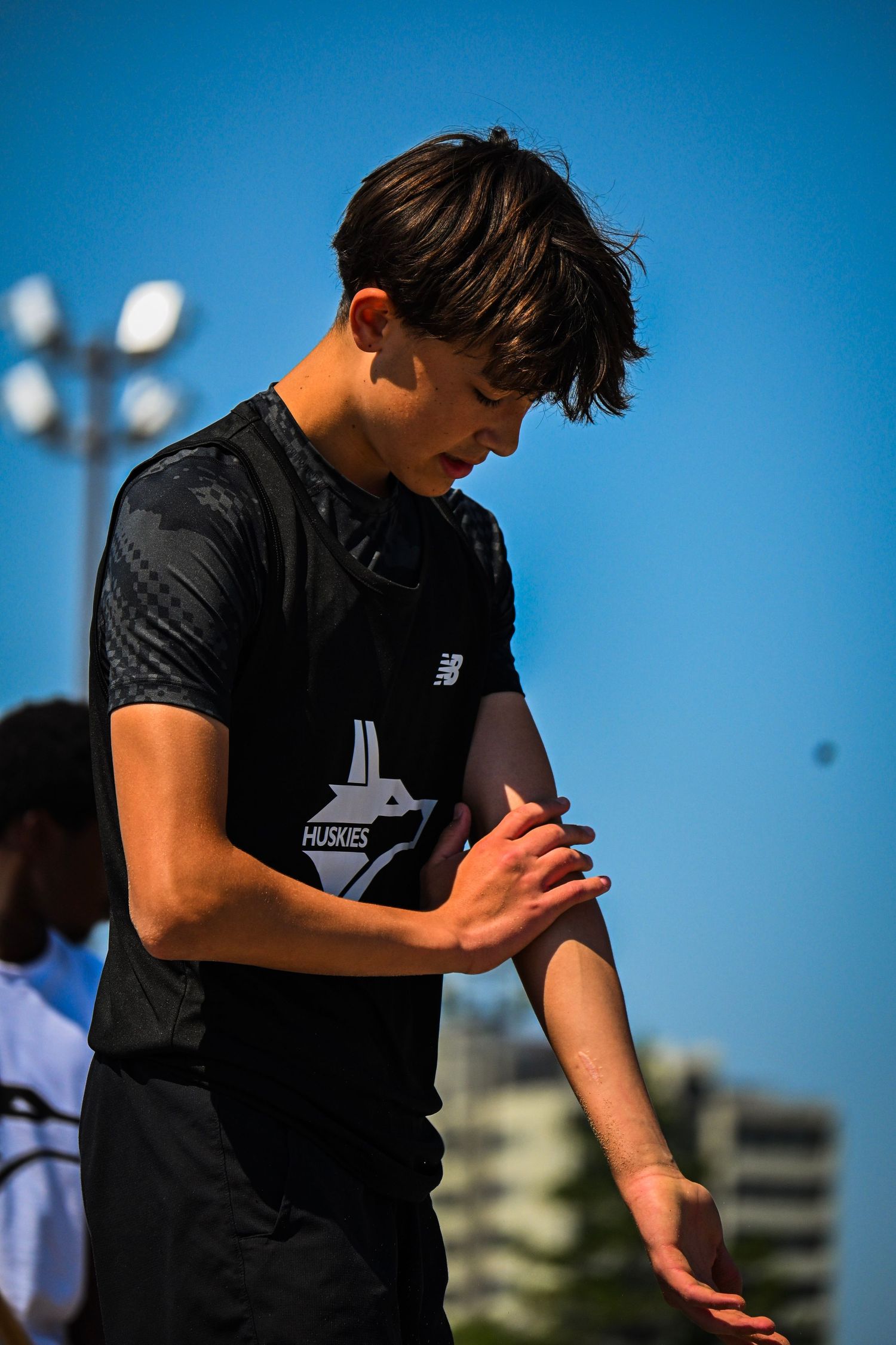 A young athlete standing outdoors looking down while adjusting his black shirt sleeve.