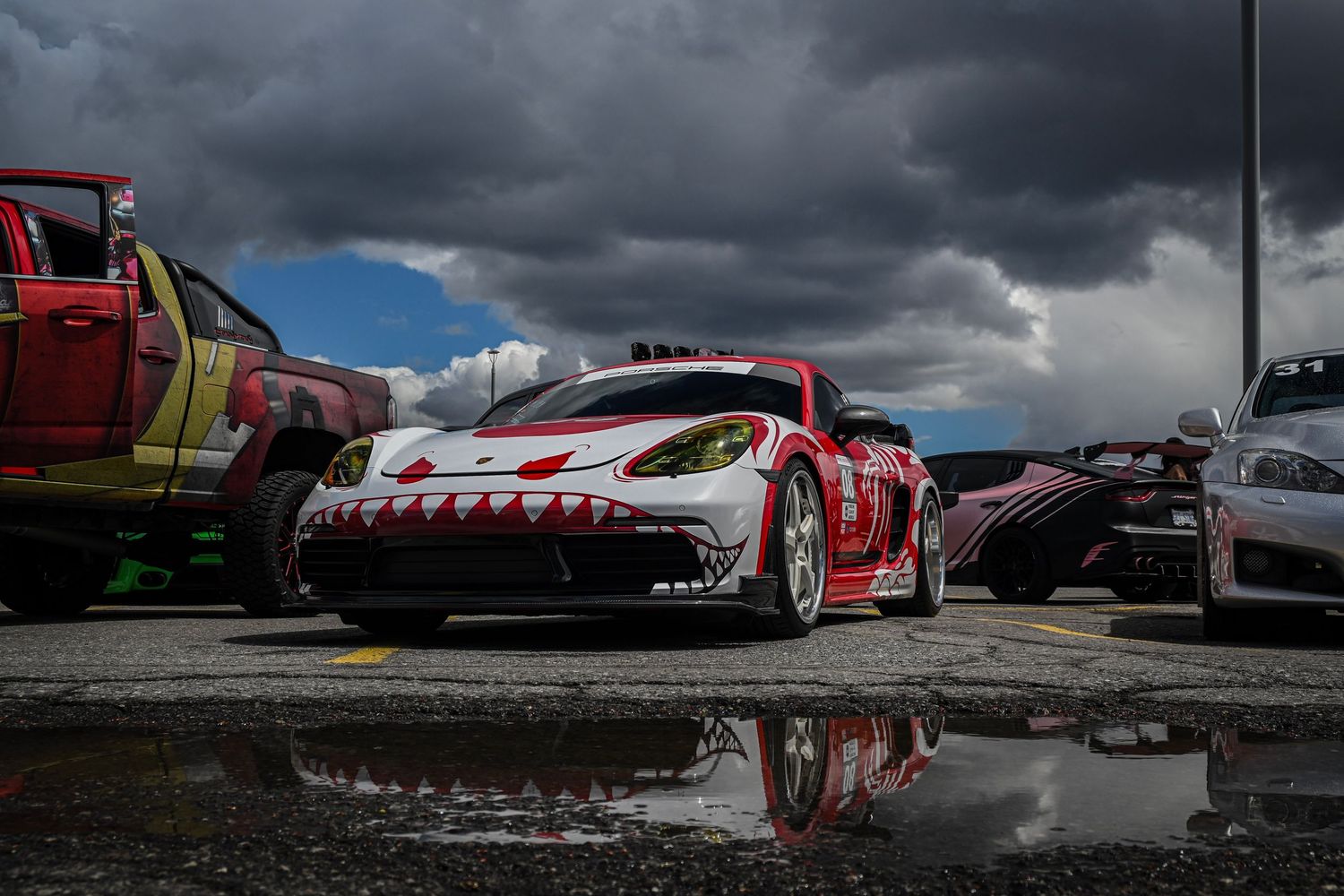 Ground-level shot of a modified red and white sports car parked outdoors with its reflection in a puddle under a dramatic, cloudy sky.