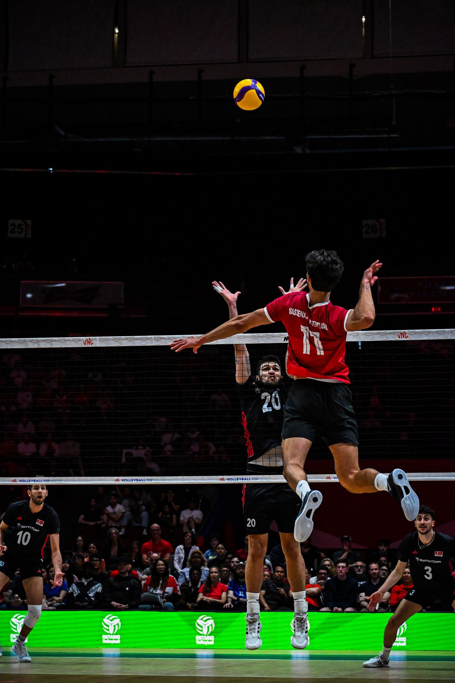 Action shot of a volleyball player in a red jersey leaping high in the air to strike the ball during an indoor match.