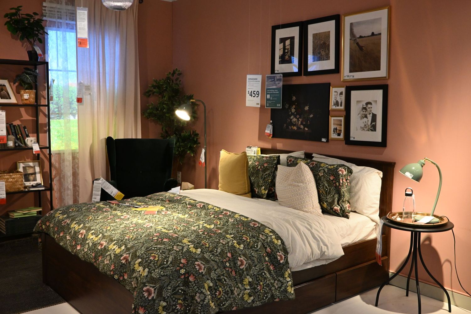 A warmly lit bedroom interior featuring a made bed with a green patterned duvet, wall art frames, and a side table.