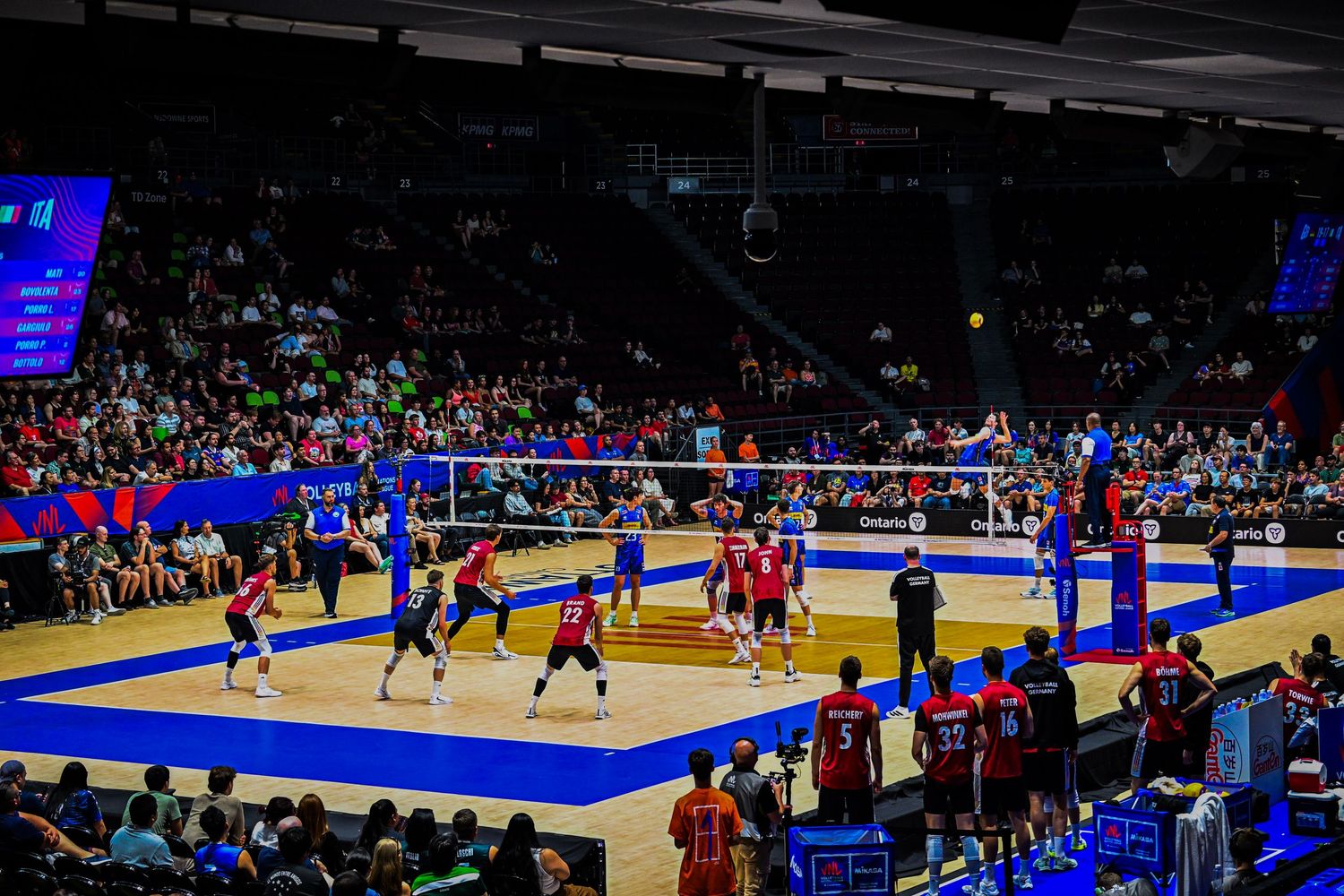 A wide view of an indoor volleyball match in a large arena packed with spectators.