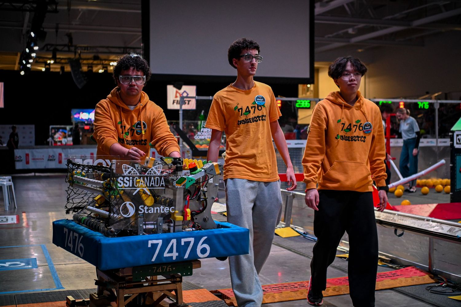 Three robotics team members in yellow sweatshirts walking alongside competition robot 7476 on a transport cart.