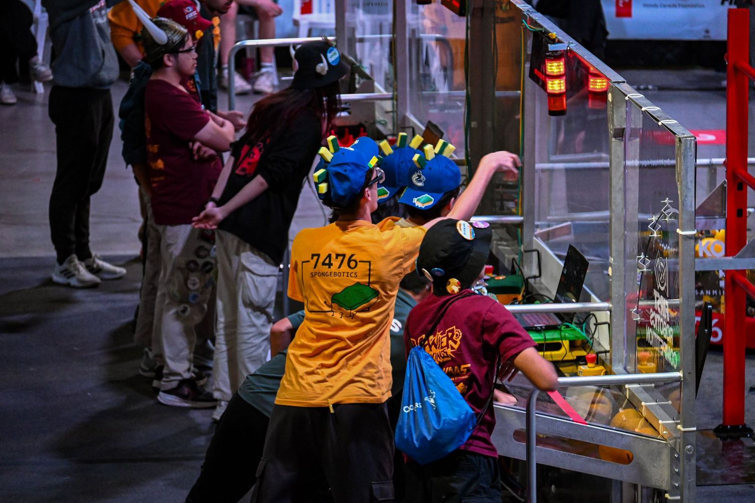Team members working on a competition robot behind a protective clear barrier in an arena.