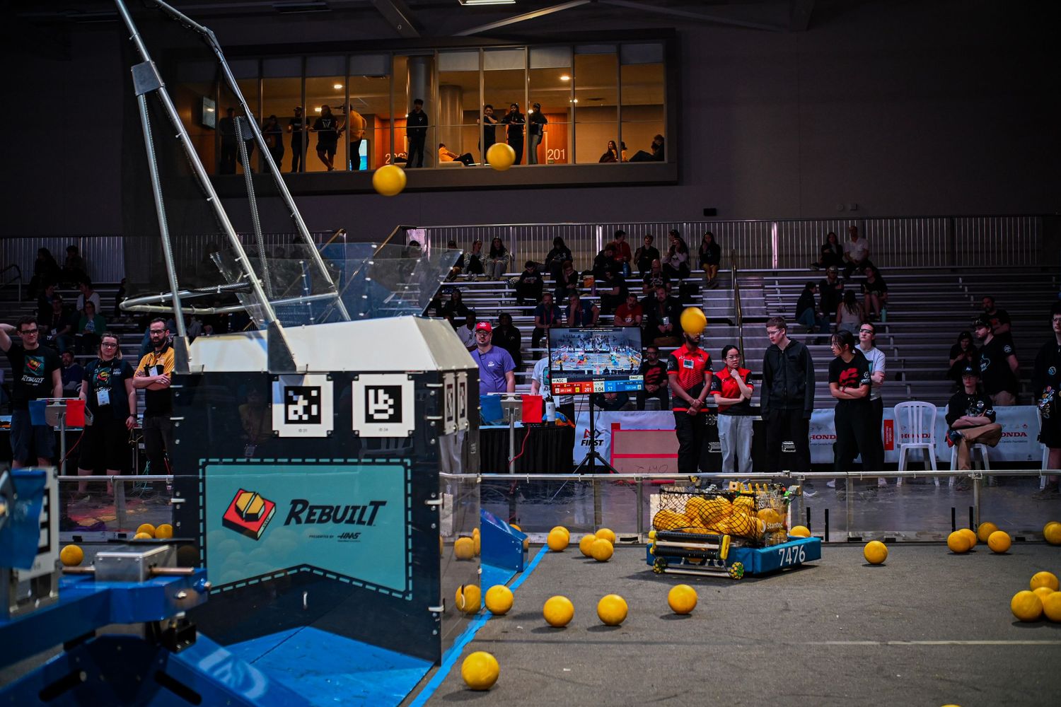 A custom robot on an indoor competition field surrounded by small yellow balls and spectators.