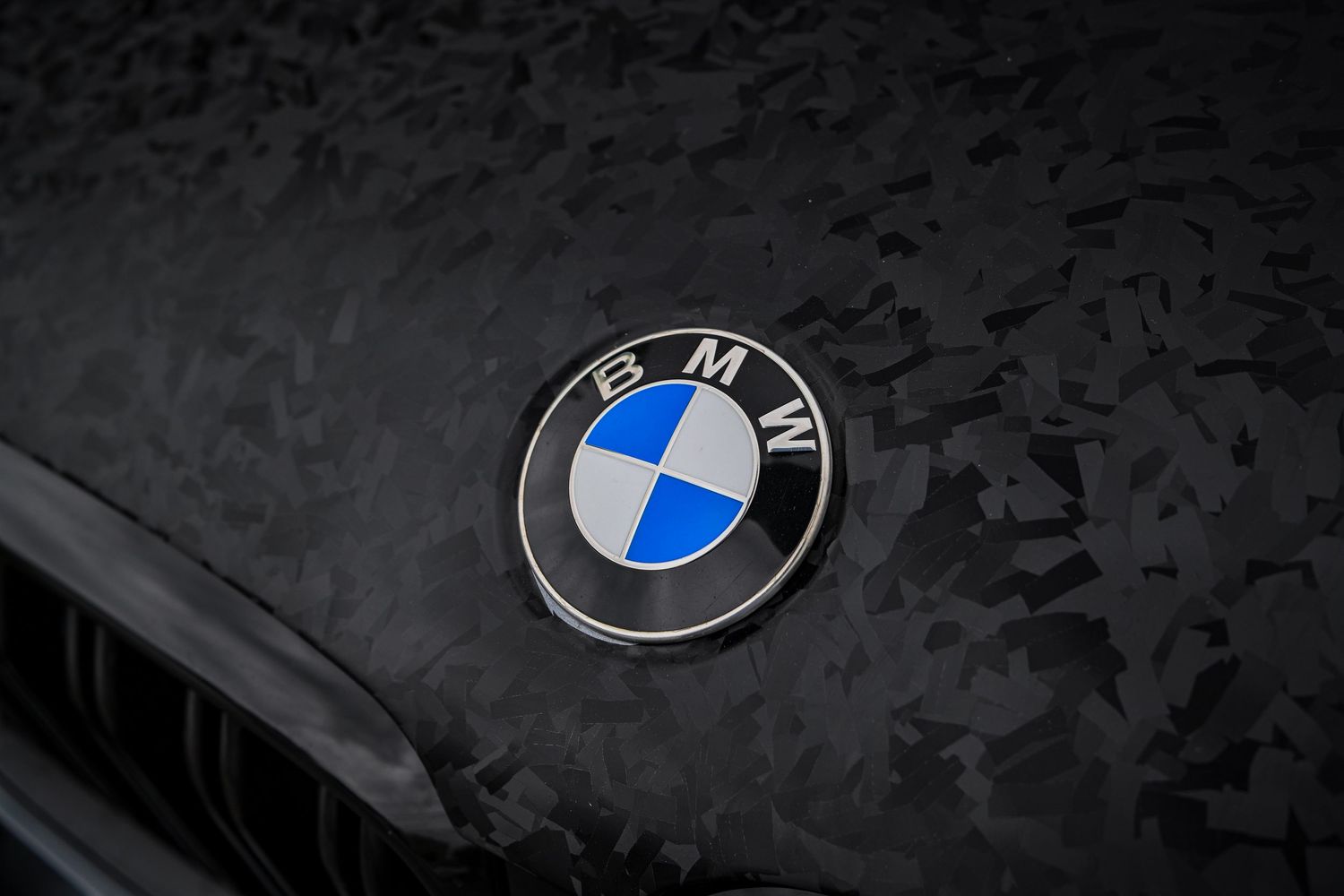 Close-up of a BMW roundel logo emblem mounted on a textured, forged carbon fiber hood.