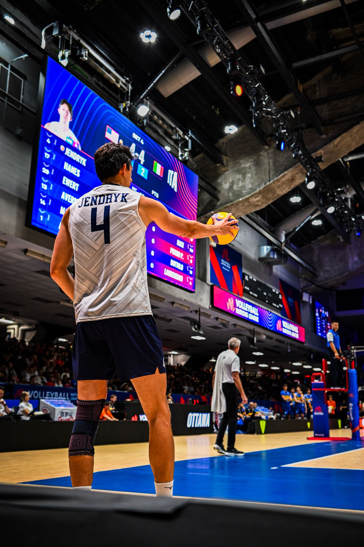 Full-length view from behind of a volleyball player in a white uniform standing on the court facing the scoreboard.