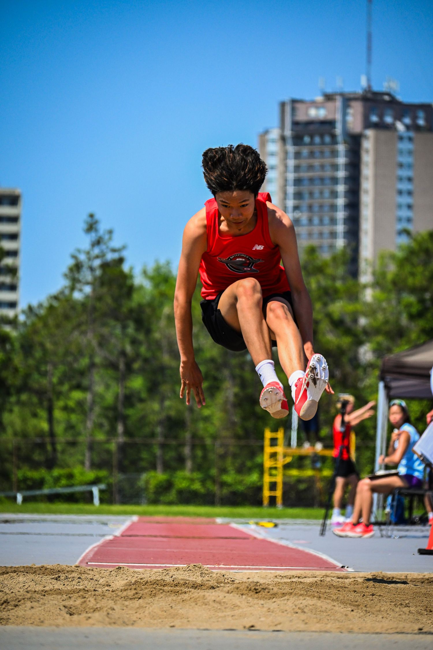 A long jumper in mid-flight with knees tucked, preparing to land in the sandpit.