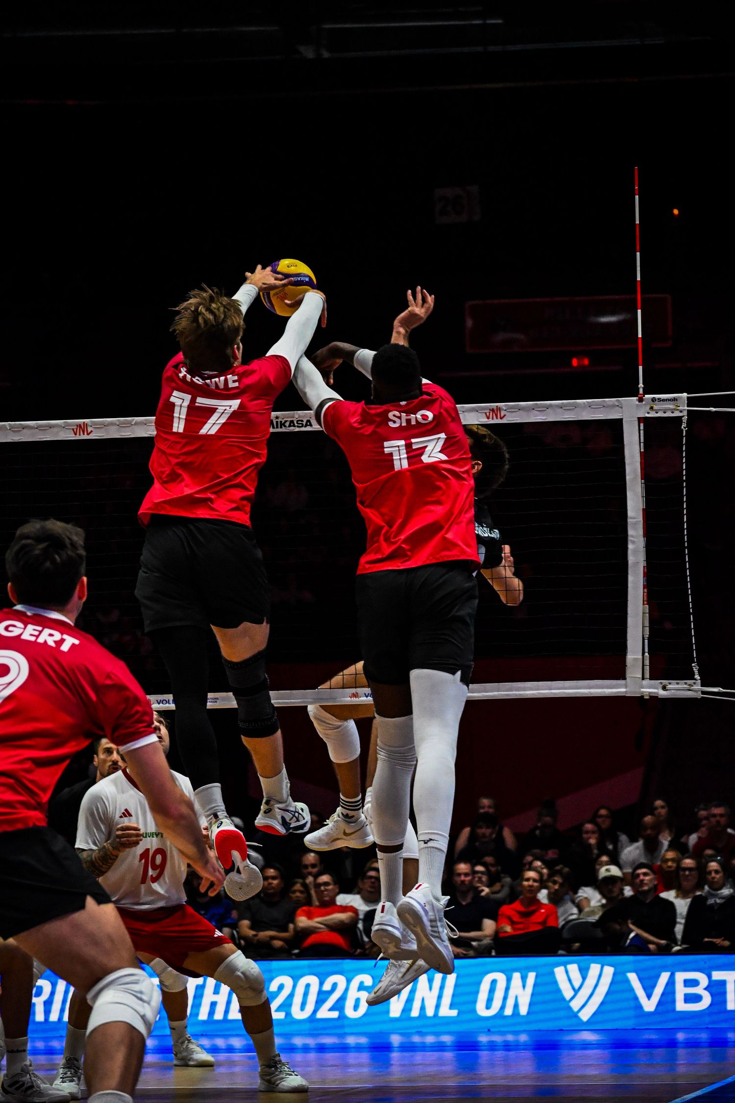 Two volleyball blockers in red jerseys leap together at the net to stop an opponent's attack.