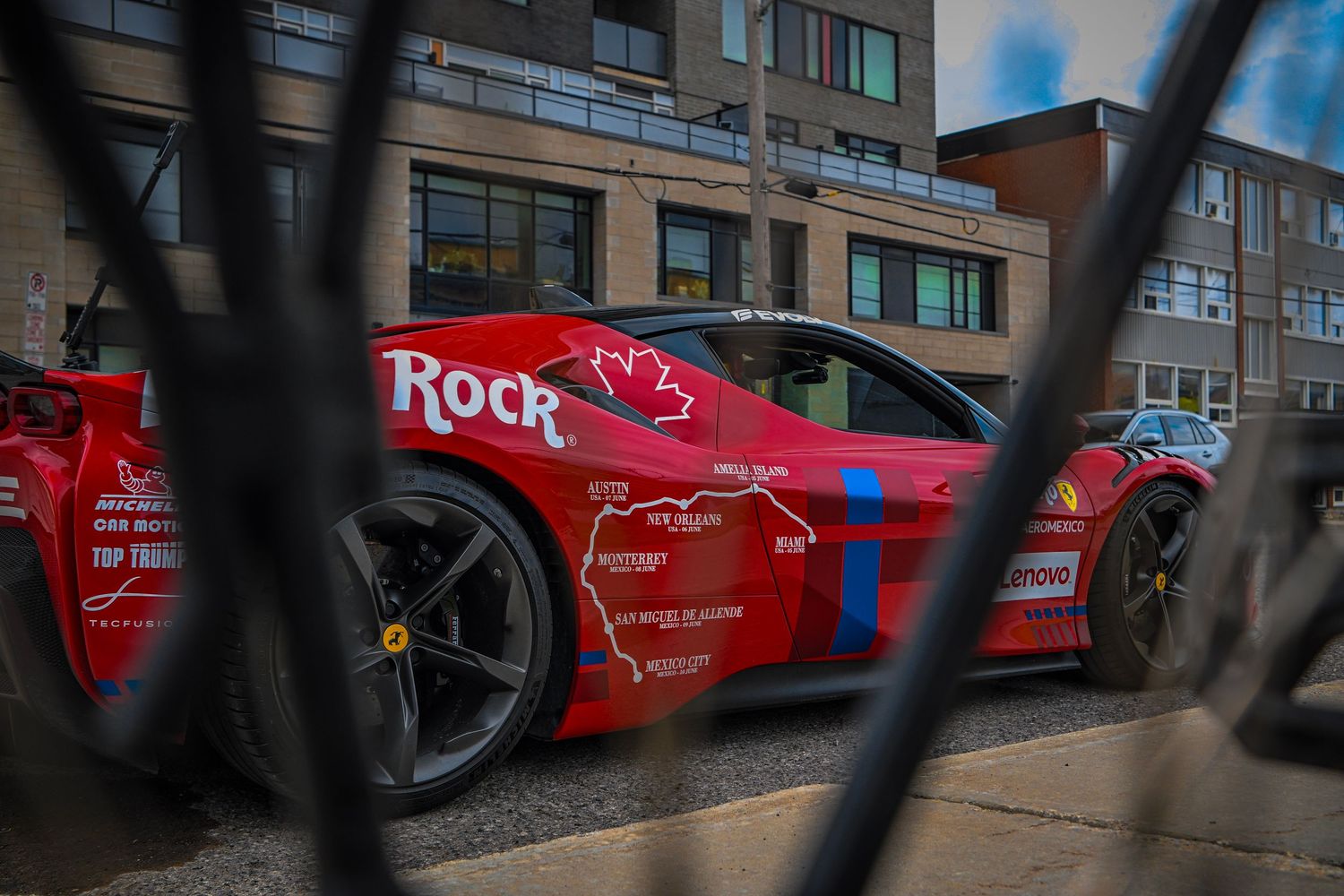 Red Ferrari race car with blue accents and sponsor graphics parked behind a metal chain-link fence on a city street.