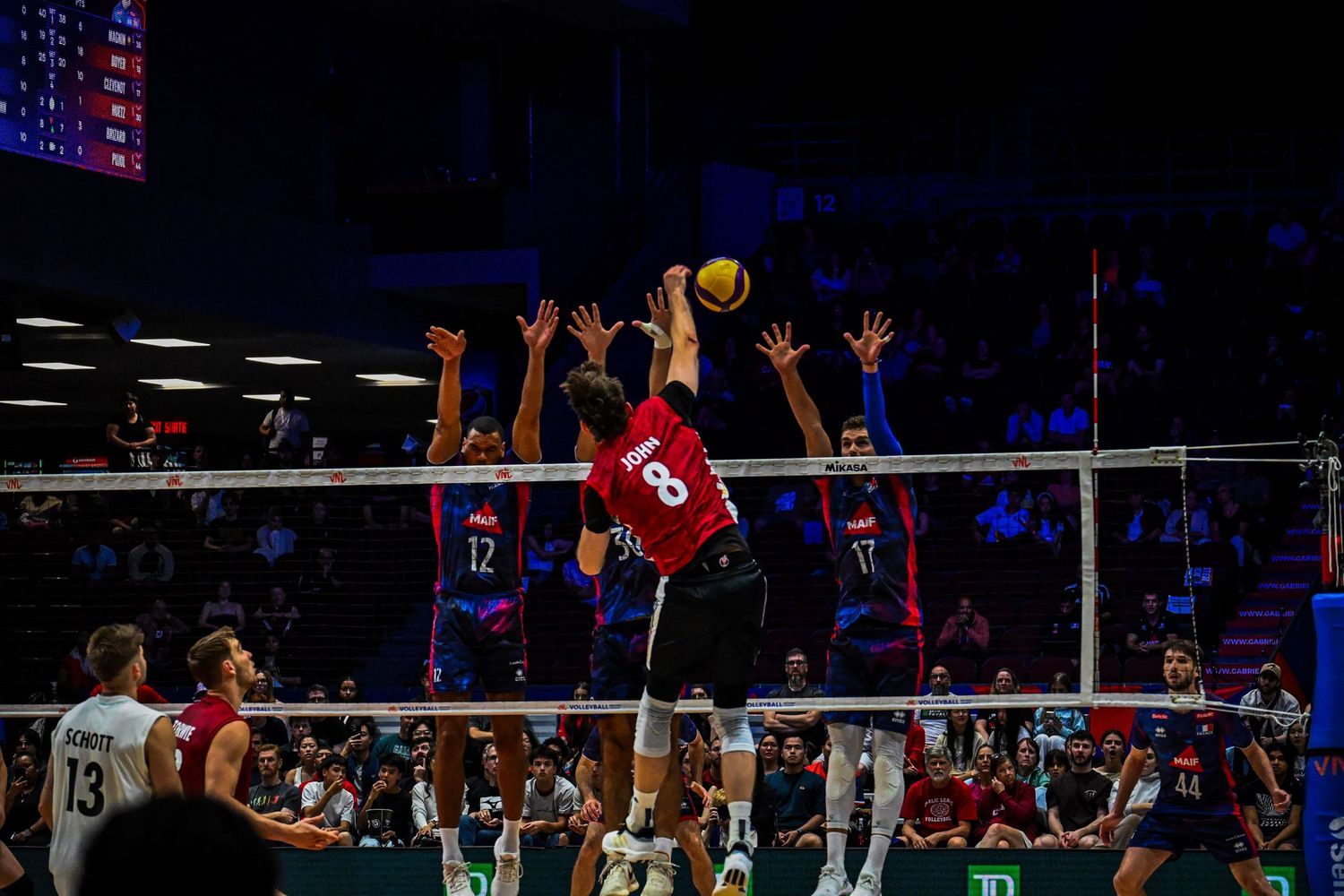 Volleyball player wearing number 8 jumps high at the net to hit the ball over multiple blocking defenders.
