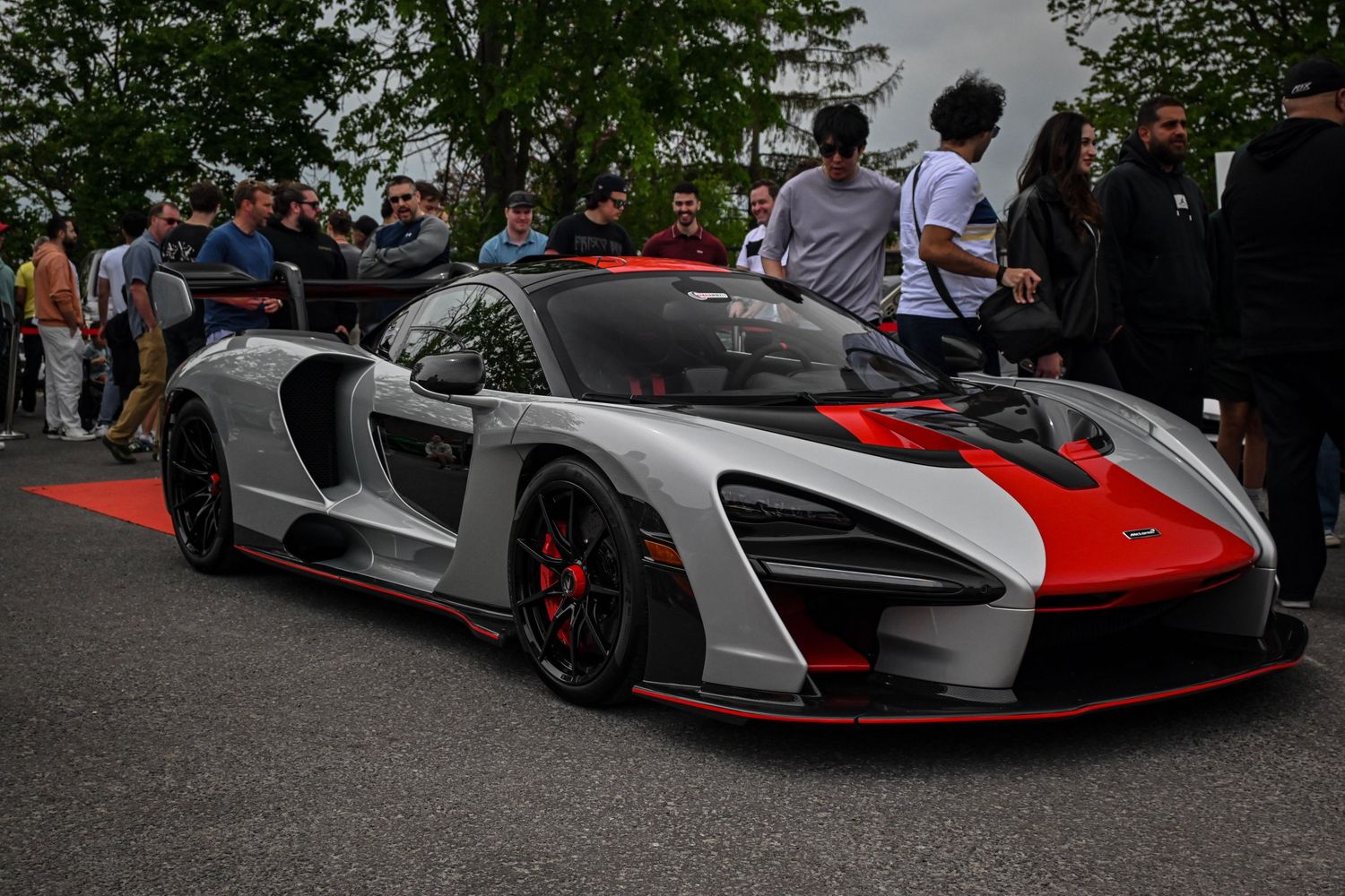 A silver and orange McLaren Senna supercar parked outdoors surrounded by a crowd of spectators.