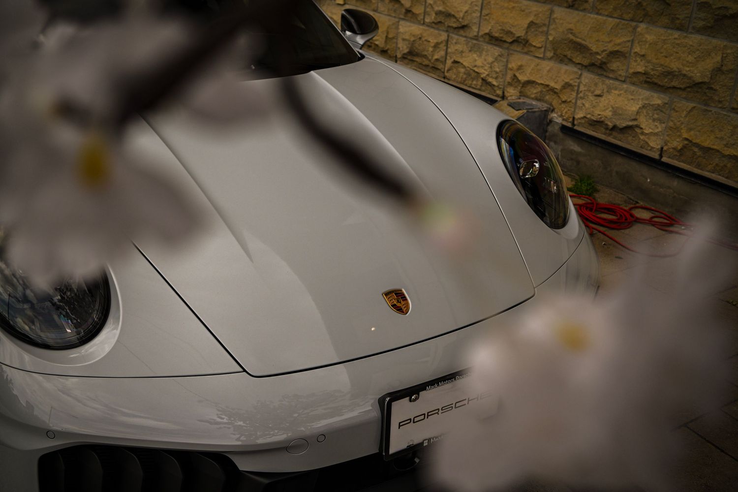 High-angle close-up of a white Porsche sports car front hood and round headlights, partially framed by blurred foreground elements.