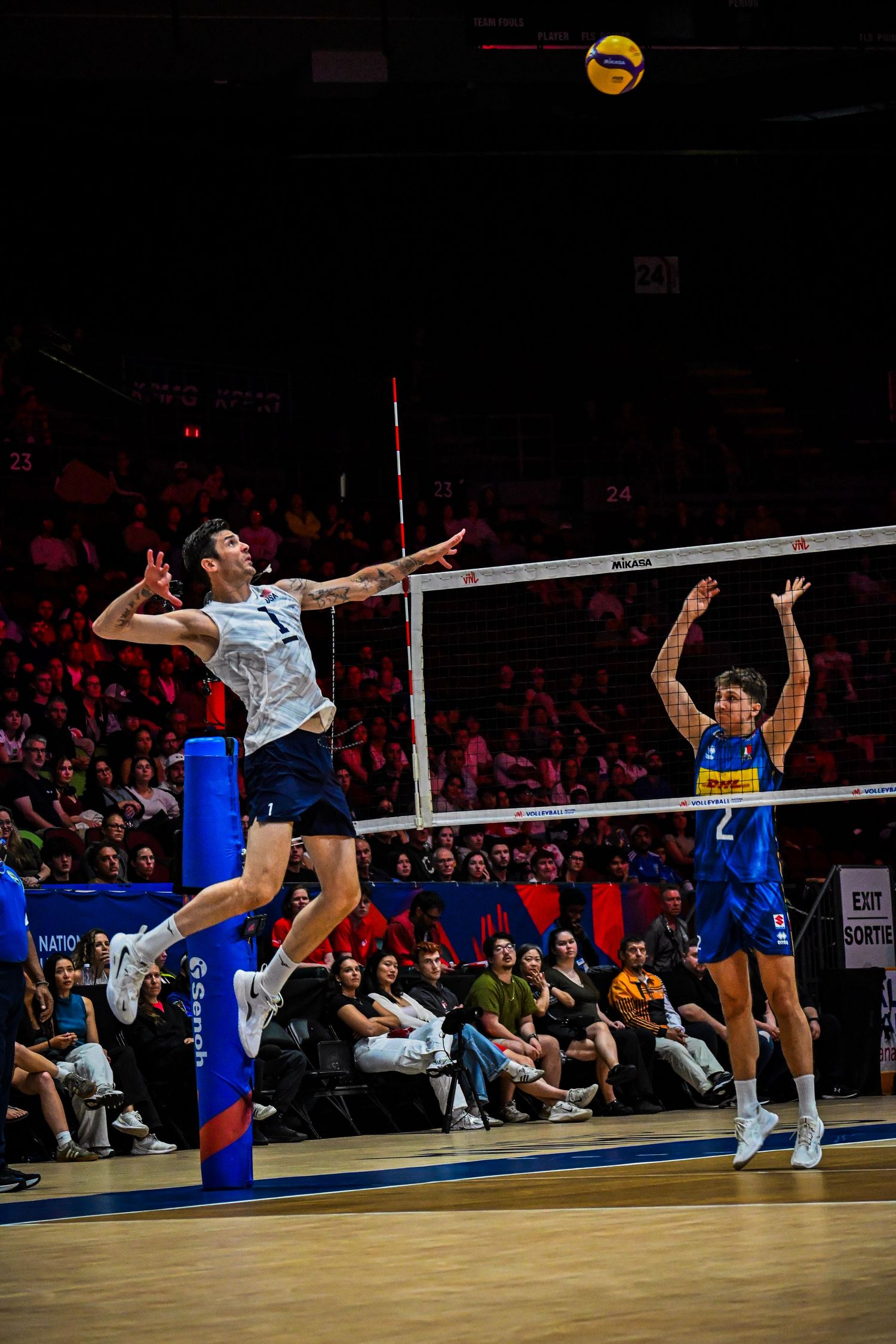 Volleyball player in a white uniform jumping high at the net to spike the ball against a defender in a blue uniform.