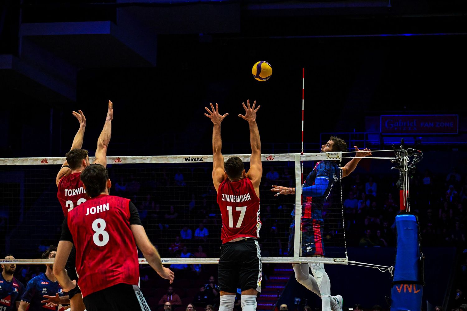 Multi-player action at the net as blockers number 8 and 17 jump to defend against an opponent spiking a volleyball.