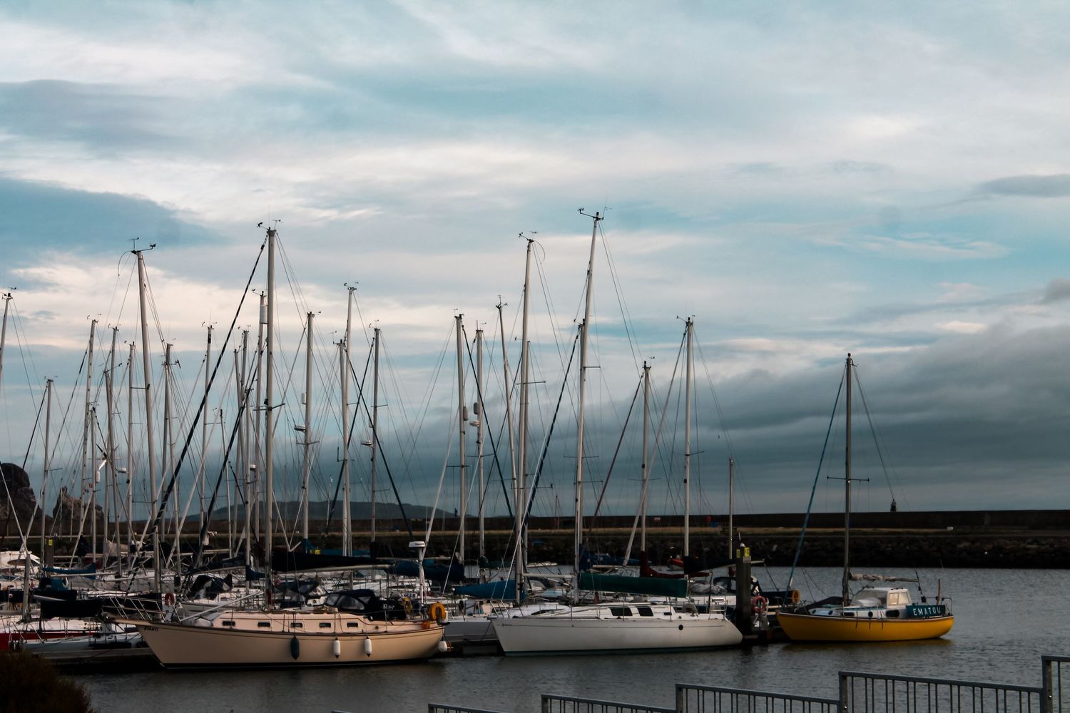 Sailboats with tall masts docked in a marina harbor under a cloudy sky, reflecting calm waters at dusk.