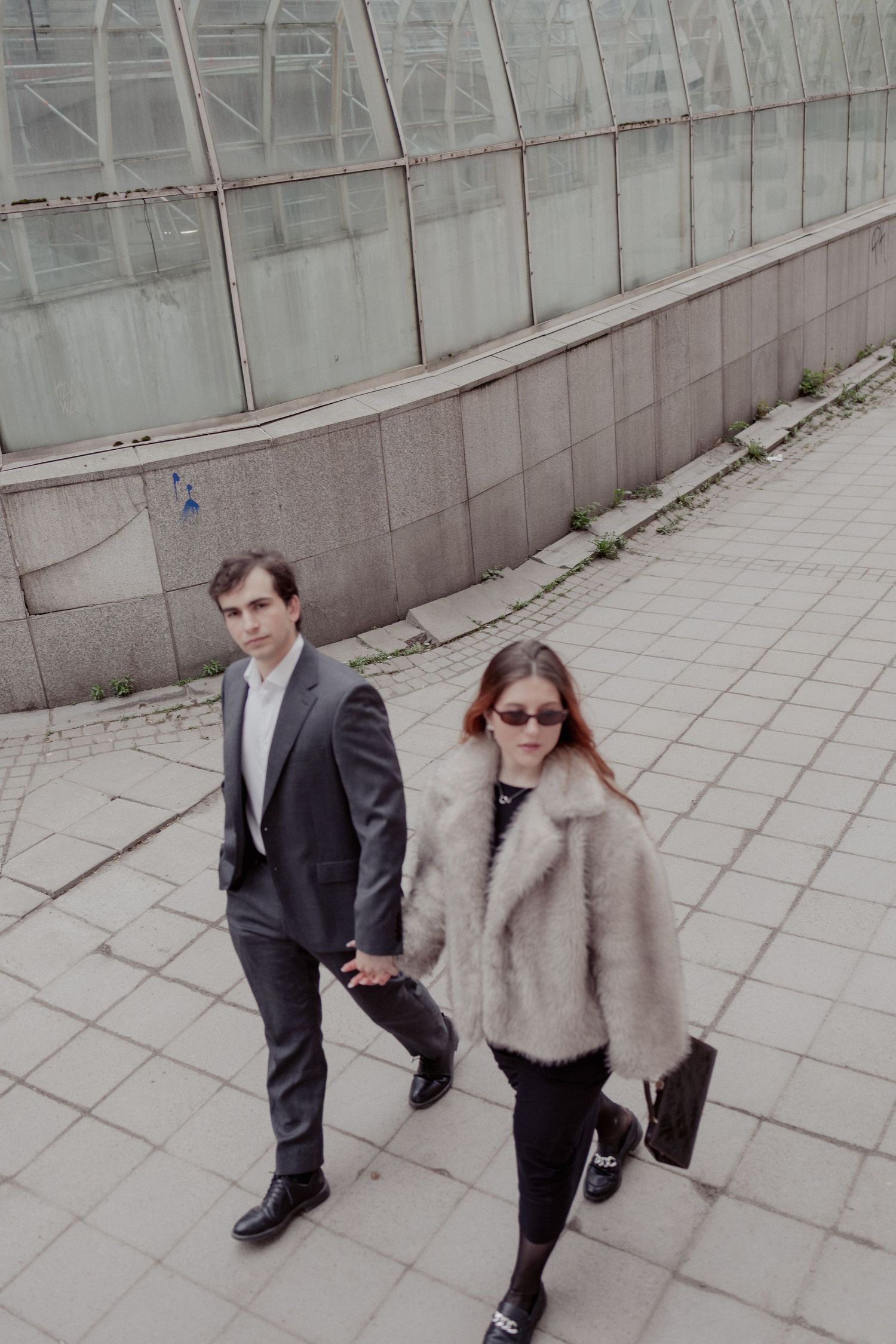 A stylish couple walks hand in hand on a city sidewalk, man in suit and woman in fur coat with sunglasses.