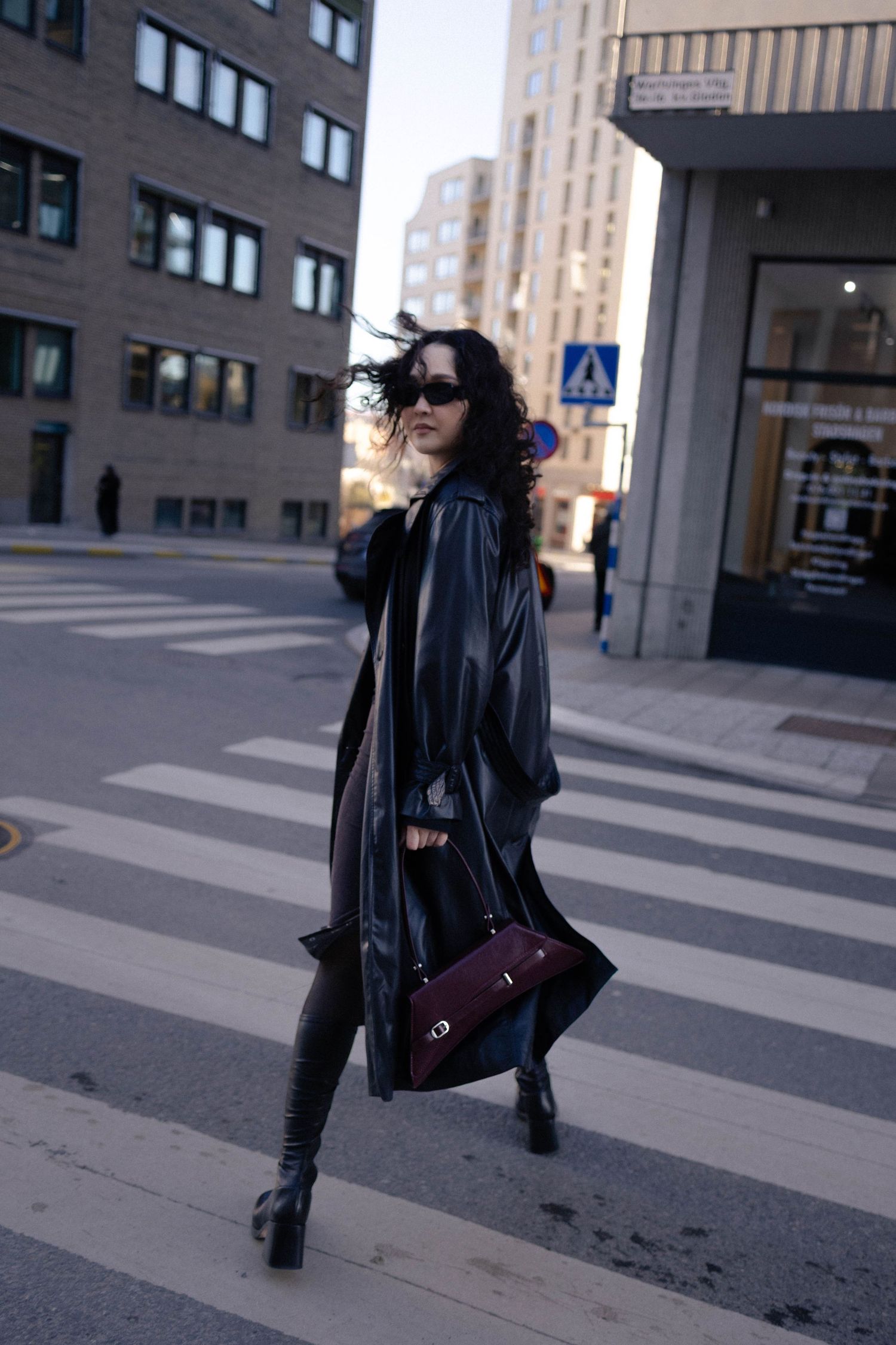 Woman in black leather coat crossing street in urban city environment.