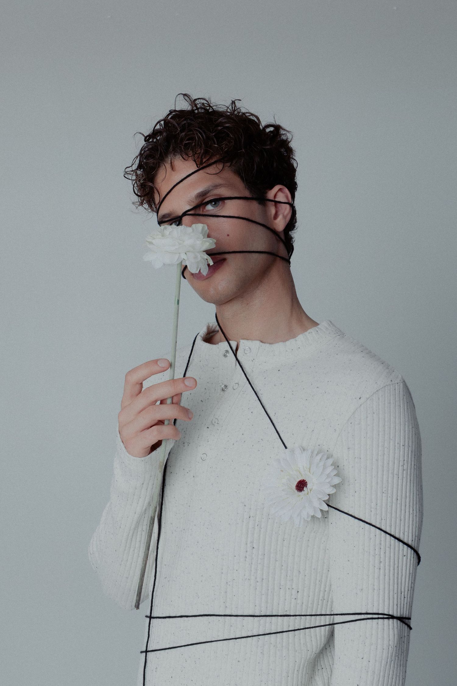 Young man in white knit sweater wrapped in black wire holding white flowers near his face.